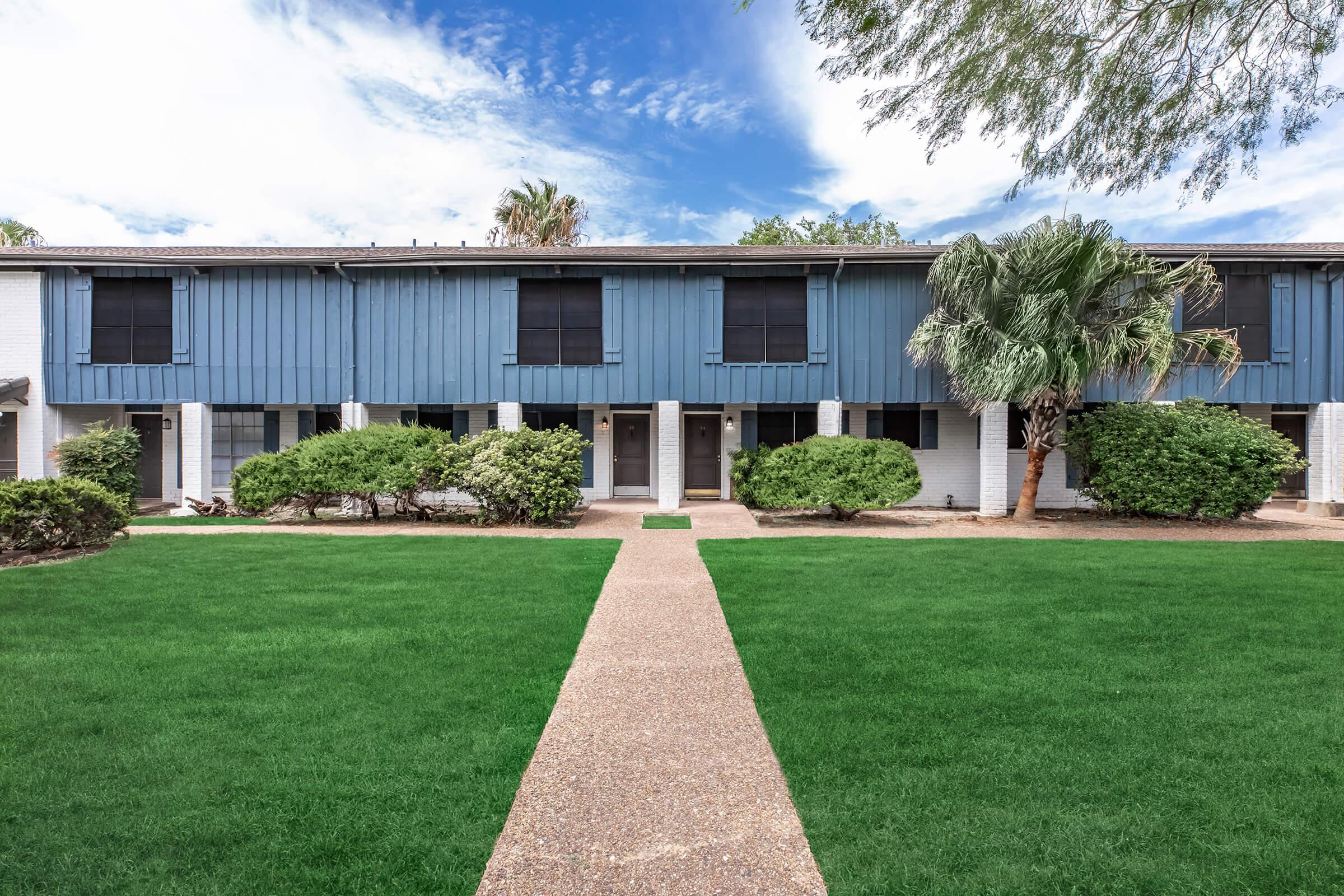 A well-maintained residential building featuring blue and white exterior, with neatly trimmed bushes and palm trees. A wide, paved walkway leads up to the entrance, surrounded by lush green grass under a partly cloudy sky.