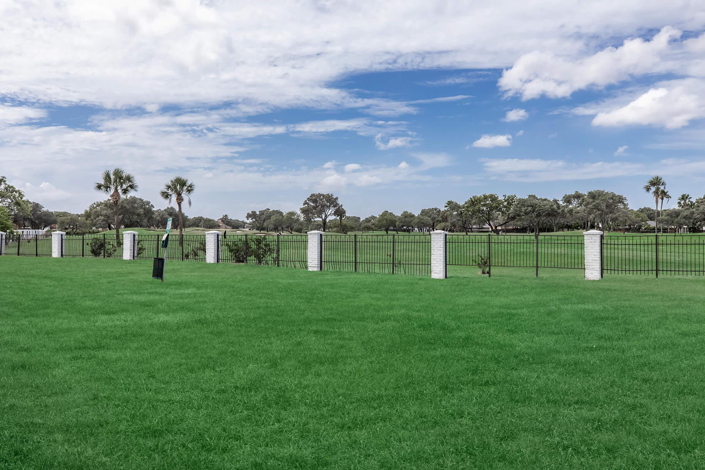 A lush green lawn stretches across the foreground, bordered by a black metal fence with white stone pillars. In the background, trees and a clear blue sky with scattered clouds create a serene landscape. The scene conveys a sense of openness and tranquility in a green space.