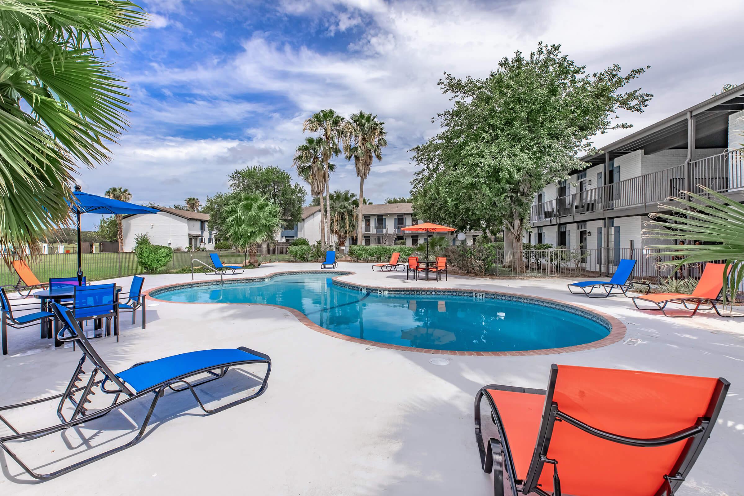 A vibrant outdoor pool area featuring a clear blue pool surrounded by lounge chairs in blue and orange, palm trees, and umbrellas. Nearby buildings provide a backdrop, with a bright sky and scattered clouds adding to the sunny atmosphere. Perfect for relaxation and leisure.