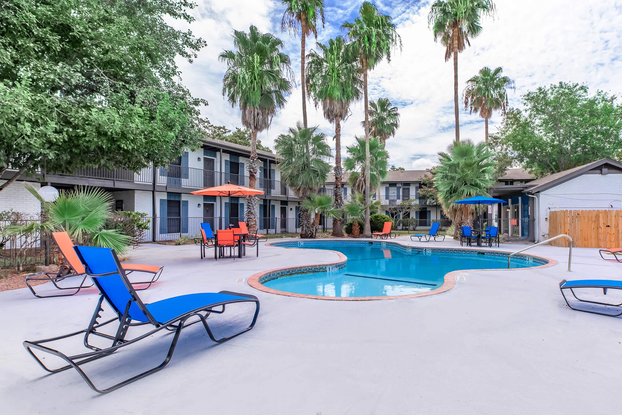 A tranquil pool area surrounded by palm trees, featuring blue and orange lounge chairs and colorful umbrellas. The pool is centered within a courtyard of an apartment complex, with well-maintained greenery and a clear sky above, creating a relaxing atmosphere.