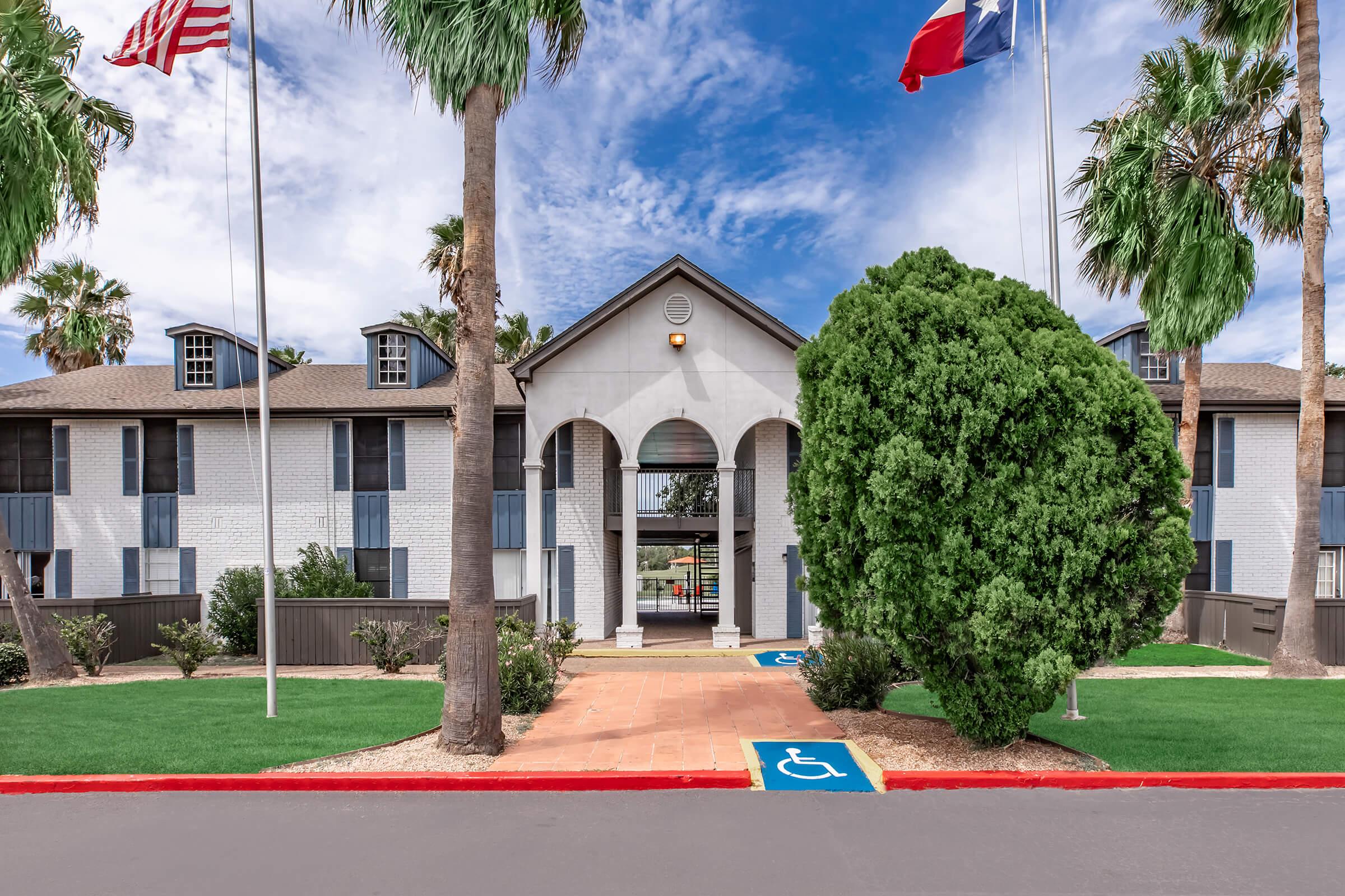 A well-maintained entrance to a building featuring palm trees, blue skies, and flags of the United States and Texas. The pathway is paved, with a visible accessible parking symbol in the foreground. The architecture includes a central archway and modern design elements typical of residential complexes.