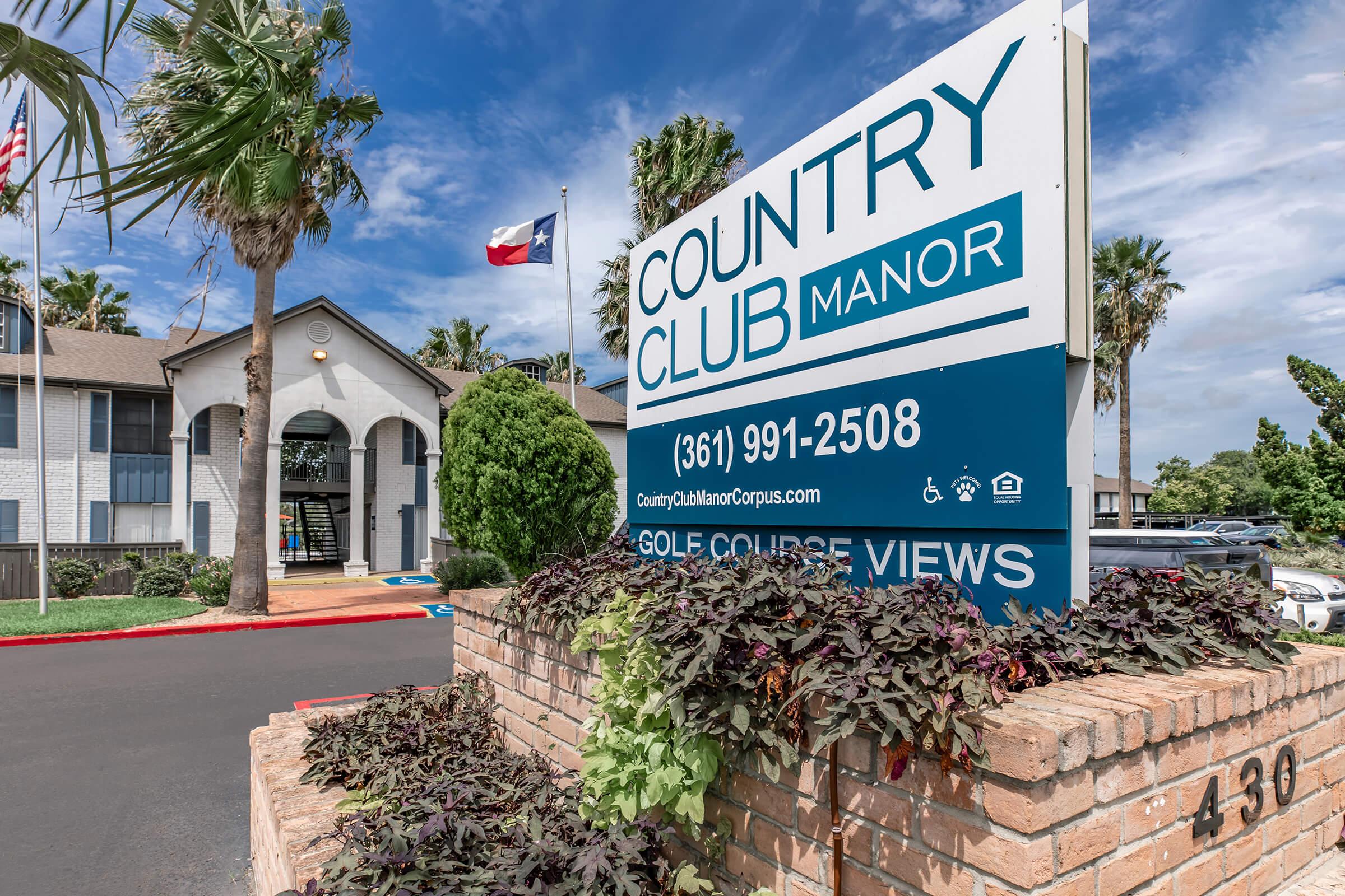 Sign for Country Club Manor featuring the name prominently. The building has palm trees and a Texas flag. The entrance is visible, with lush greenery and colorful plants in the foreground. Contact number and website are displayed on the sign.