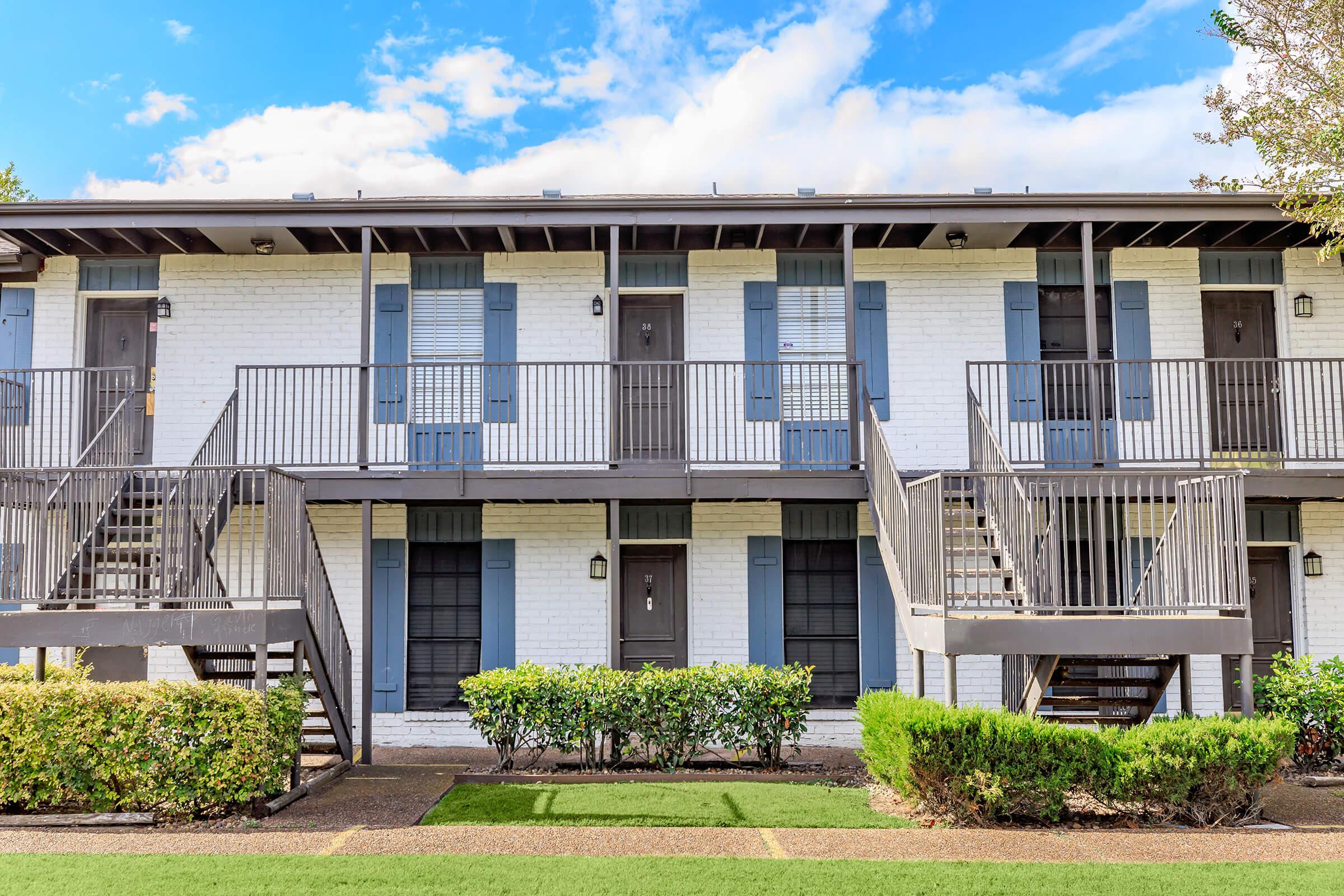 A two-story apartment building featuring a white exterior, black railings, and blue shutters. There are two sets of stairs leading to upper and lower entrances. The landscape includes well-maintained greenery in front and a clear blue sky overhead.