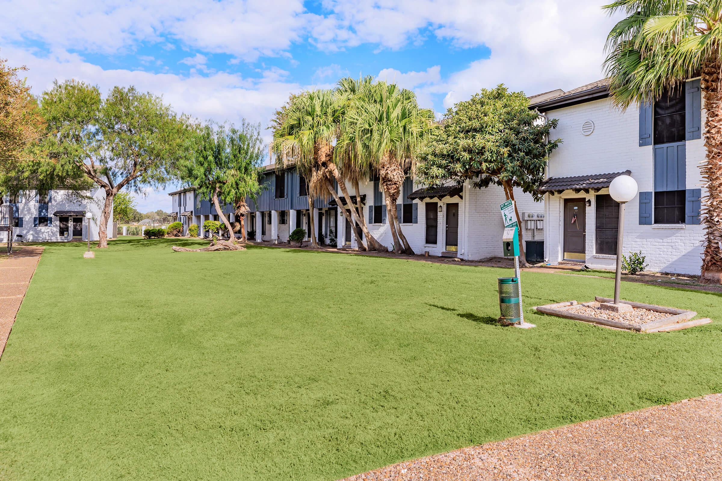 A well-maintained outdoor area featuring a lush green lawn, palm trees, and several white two-story buildings. The scene is bright and sunny with a blue sky and some scattered clouds, creating a welcoming environment in a residential setting.