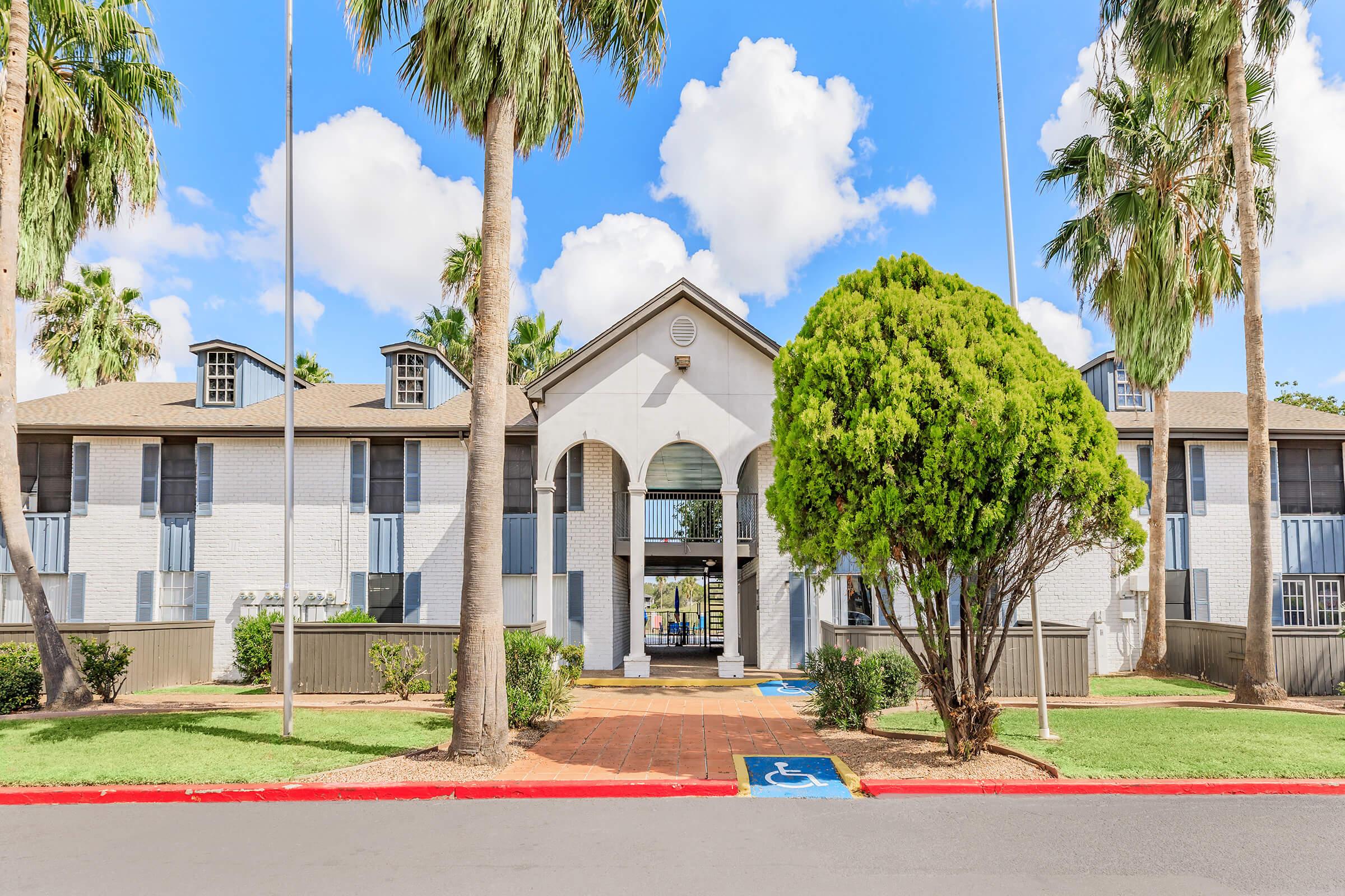 A well-maintained, two-story building with a white exterior and archway entrance, surrounded by palm trees and green landscaping. The scene features a clear blue sky and a paved path leading to the entrance, with a wheelchair-accessible parking spot visible in front.