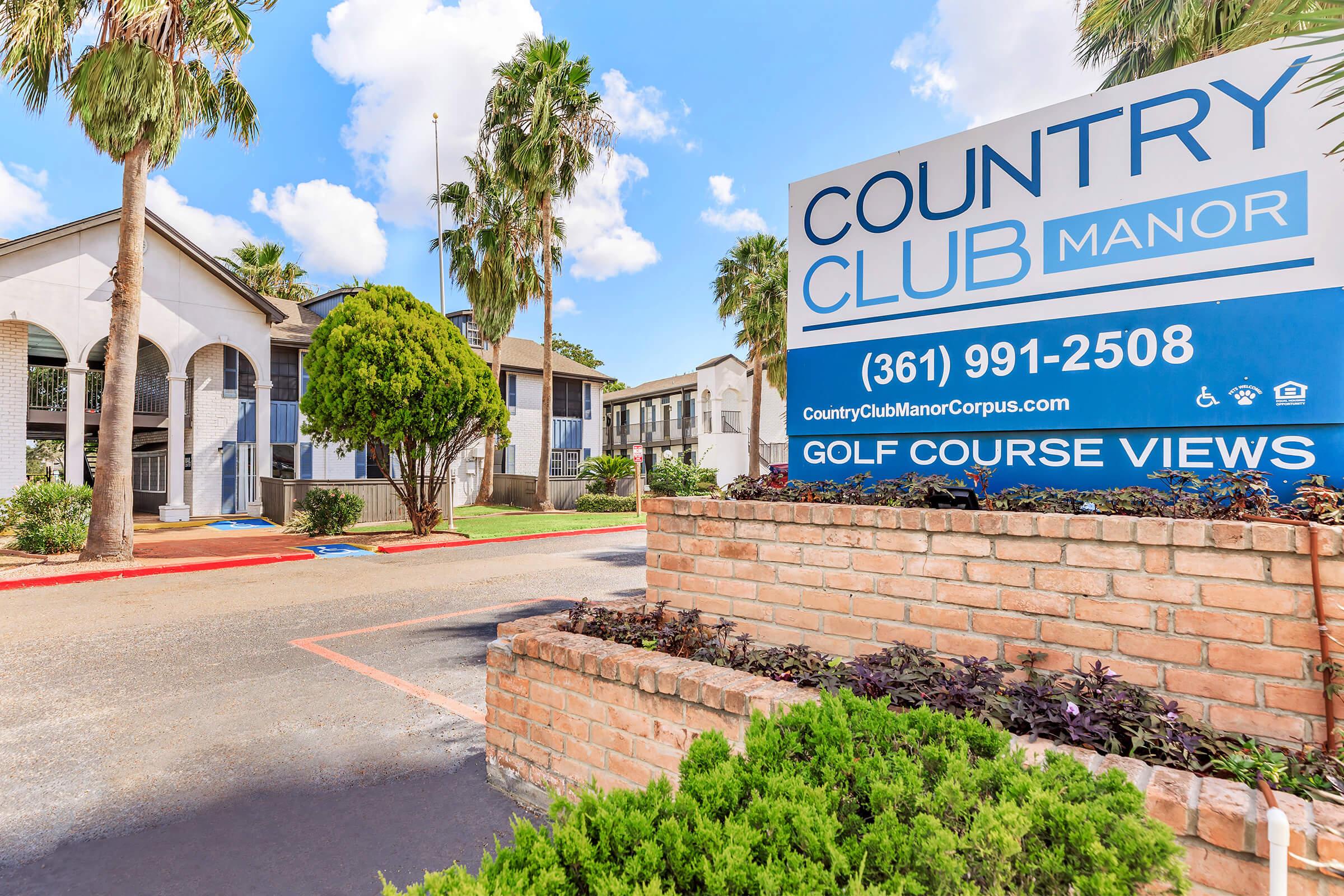 A sign for Country Club Manor featuring a blue background with white text, promotional details, and contact information. The building is visible in the background, surrounded by palm trees and a well-maintained landscape, indicating a welcoming environment near a golf course.