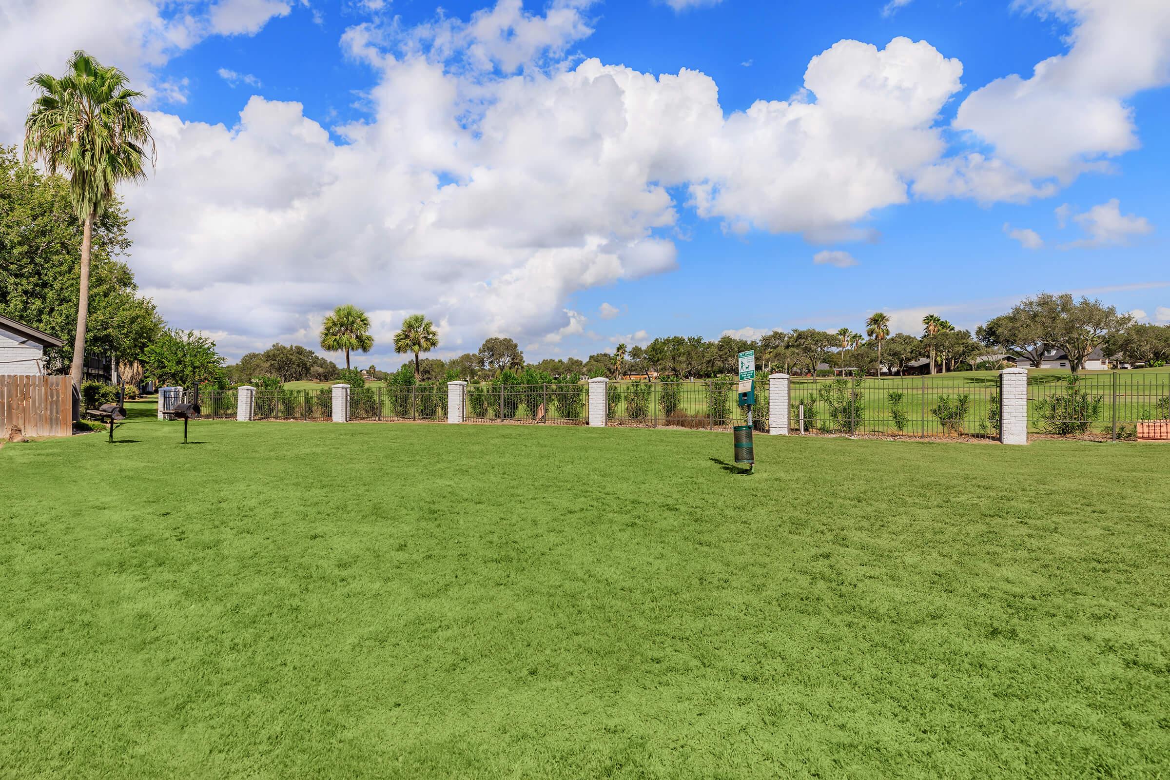 A bright green park area with freshly mowed grass, surrounded by a fence and palm trees. The sky is partly cloudy with blue patches peeking through. There are decorative stone pillars and a few benches visible in the background, creating a calm and inviting outdoor space.