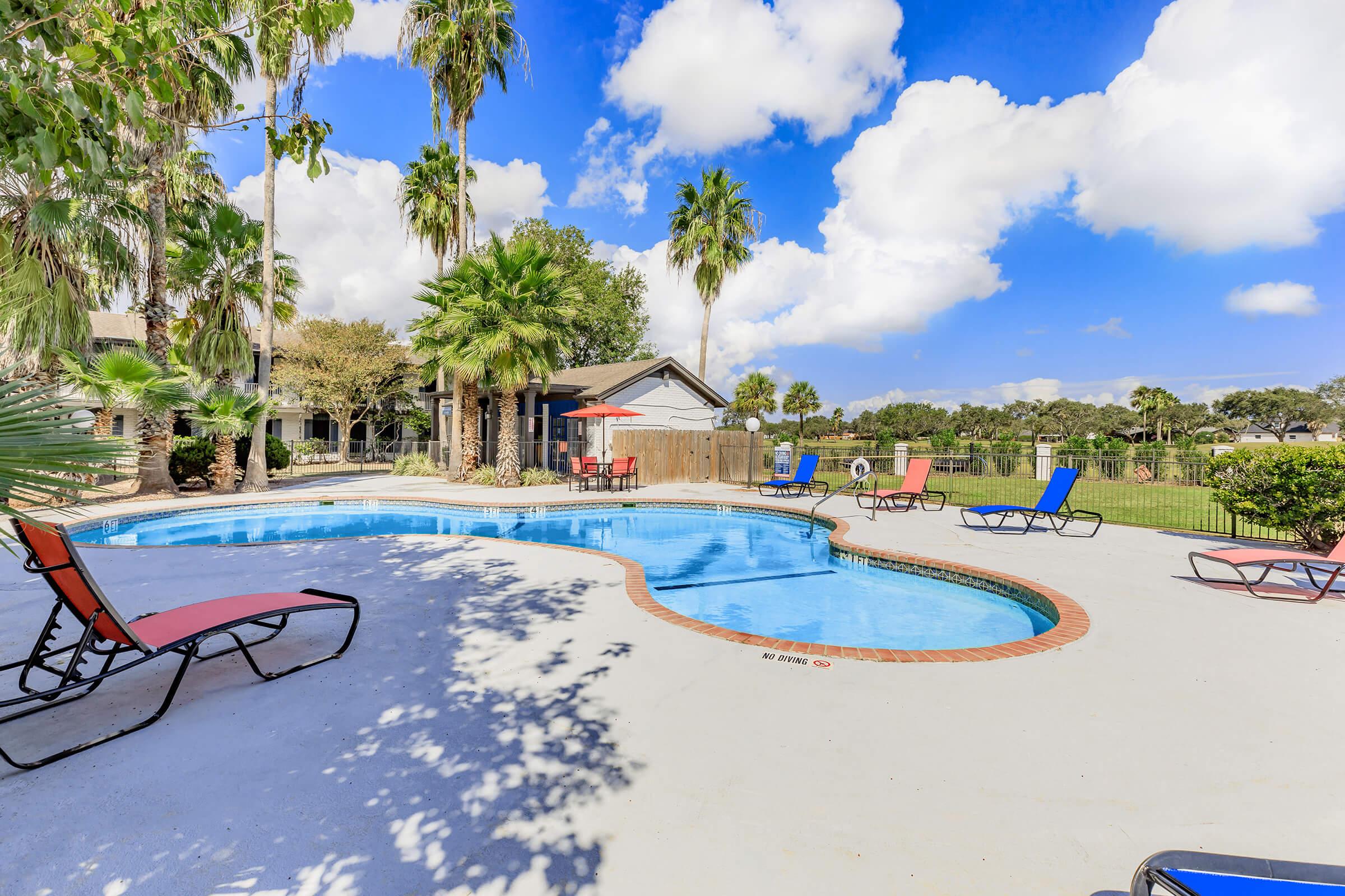A sunny outdoor pool area featuring a curved swimming pool surrounded by palm trees. Colorful lounge chairs are placed around the pool, with a patio umbrella providing shade. The sky is bright blue with fluffy white clouds, and a landscaped lawn is visible in the background.