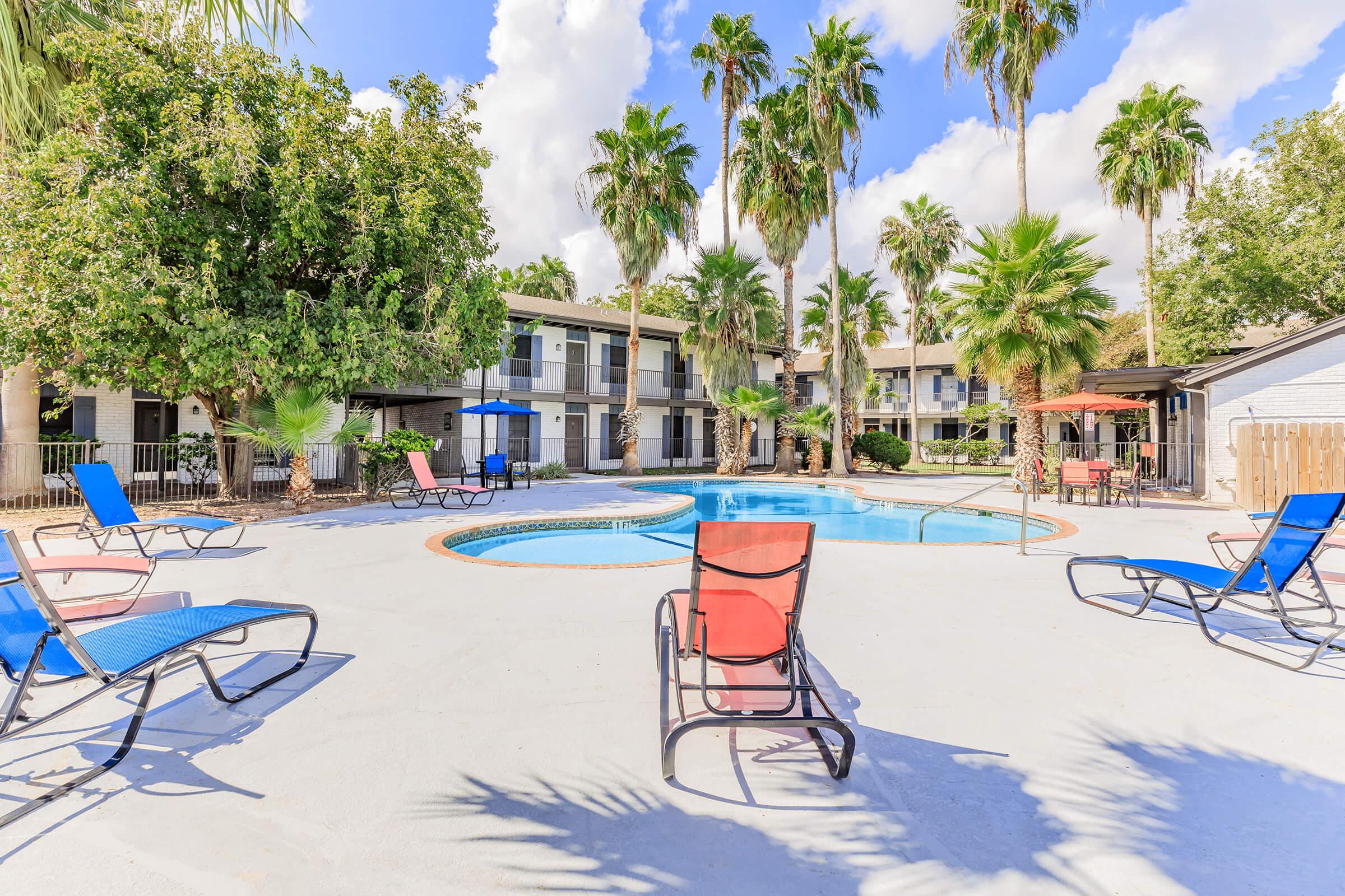 A sunny pool area featuring a circular swimming pool surrounded by palm trees. Lounge chairs in red and blue are arranged around the pool deck, creating a relaxing atmosphere. There are shaded areas with umbrellas and various greenery, set against a bright blue sky with fluffy white clouds.