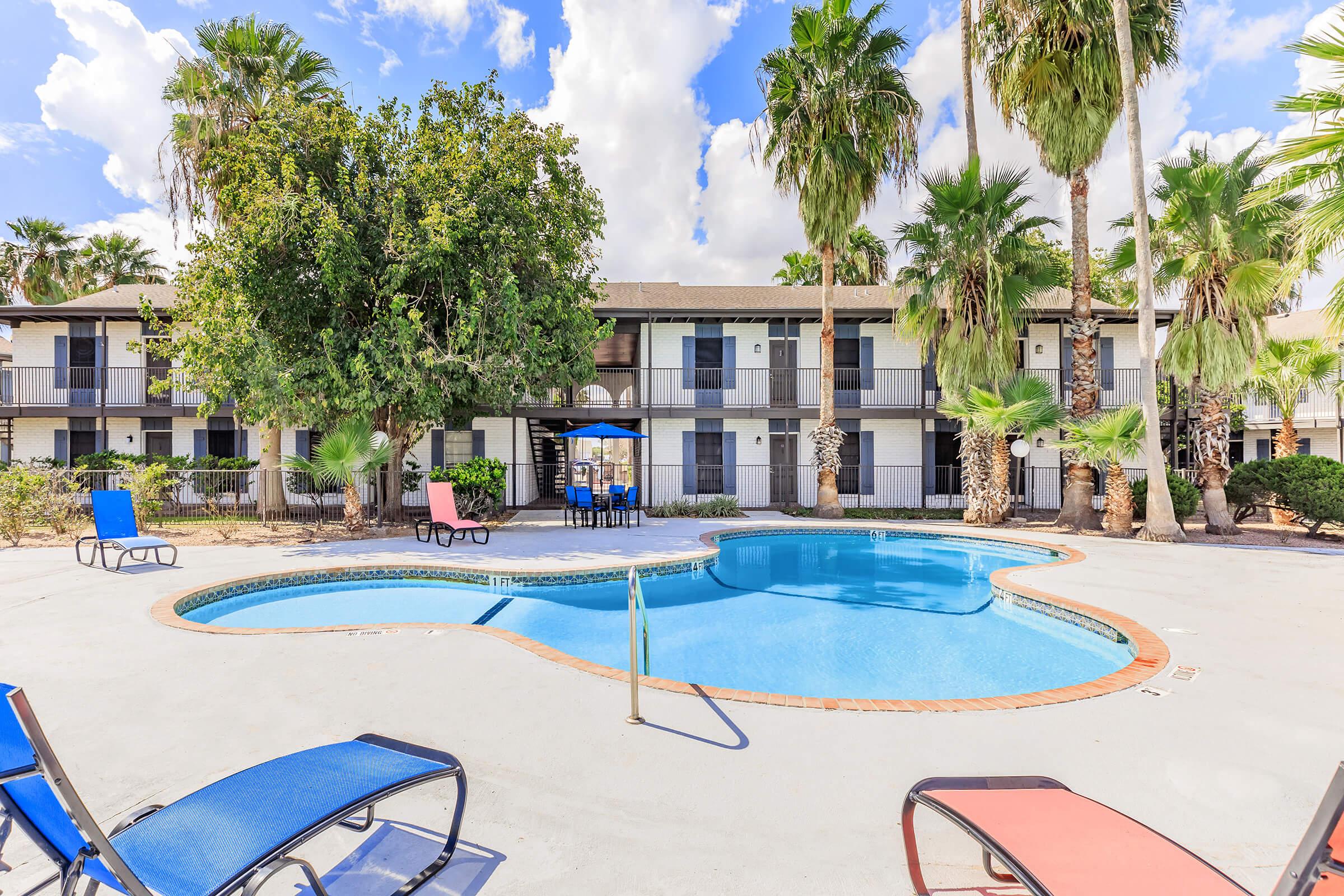 A swimming pool surrounded by lounge chairs and palm trees, with a backdrop of a two-story apartment building. The pool area features a table with umbrellas, creating a relaxed outdoor atmosphere. Bright blue skies with fluffy white clouds enhance the inviting scene.