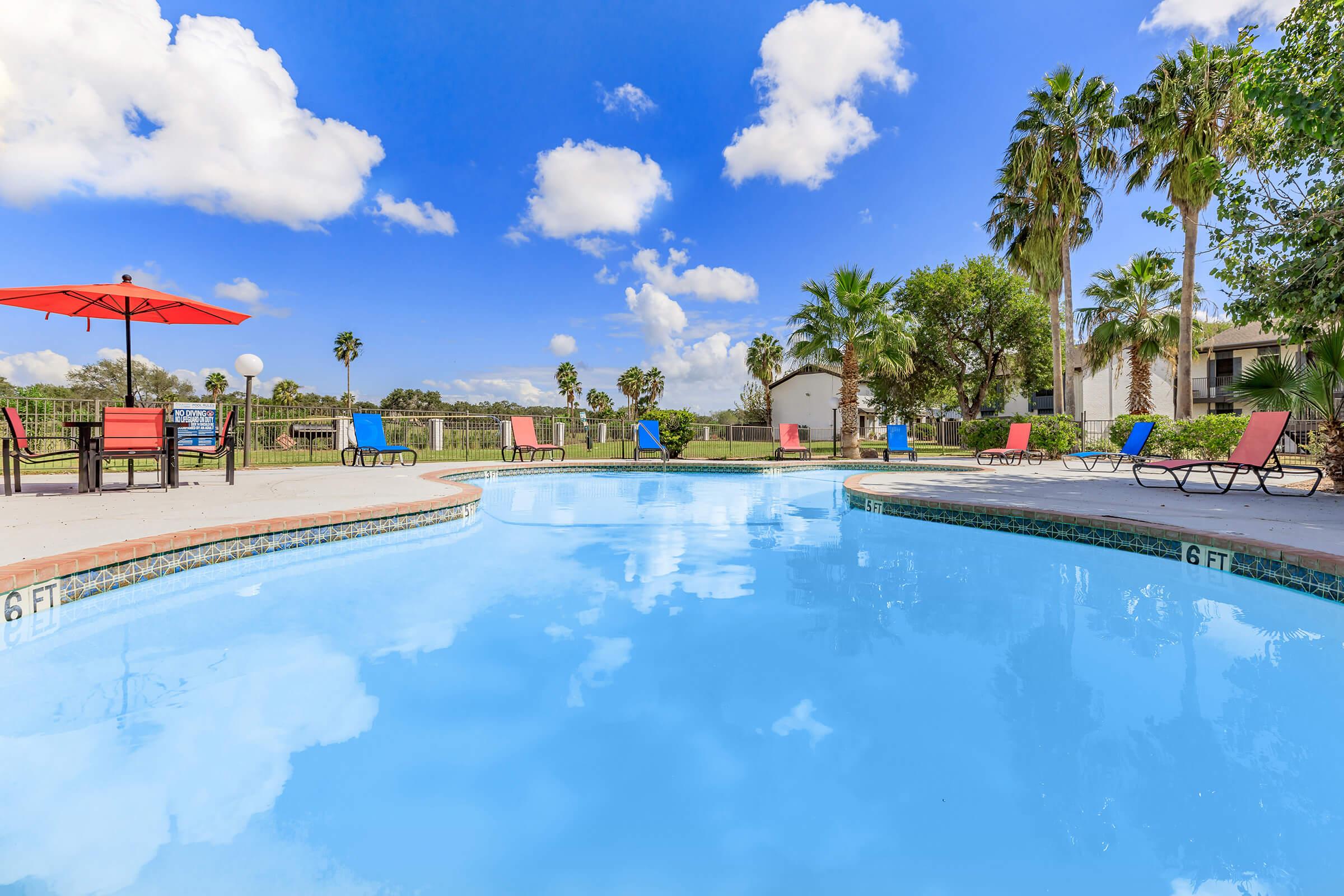 A sparkling blue swimming pool surrounded by lounge chairs in sunny weather. Palm trees and greenery line the background, while fluffy white clouds float in a bright blue sky. An umbrella provides shade over part of the pool area, creating a relaxing outdoor space.