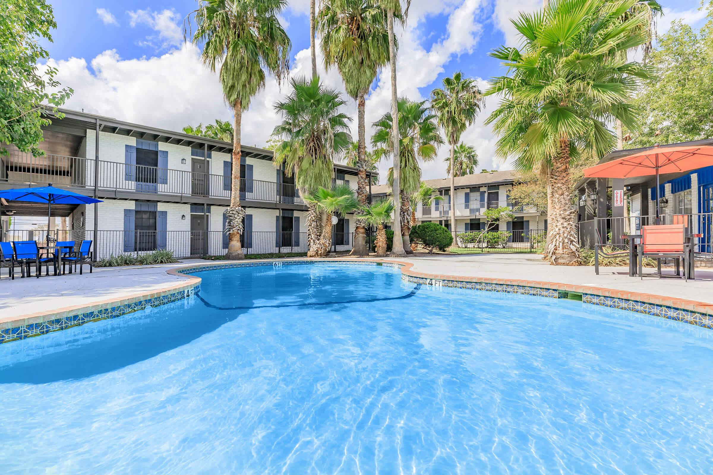 A vibrant swimming pool surrounded by palm trees and lounge chairs, with two-story buildings in the background. Bright blue skies with scattered clouds complete the sunny, inviting atmosphere. Perfect for relaxation and leisure in a tropical setting.