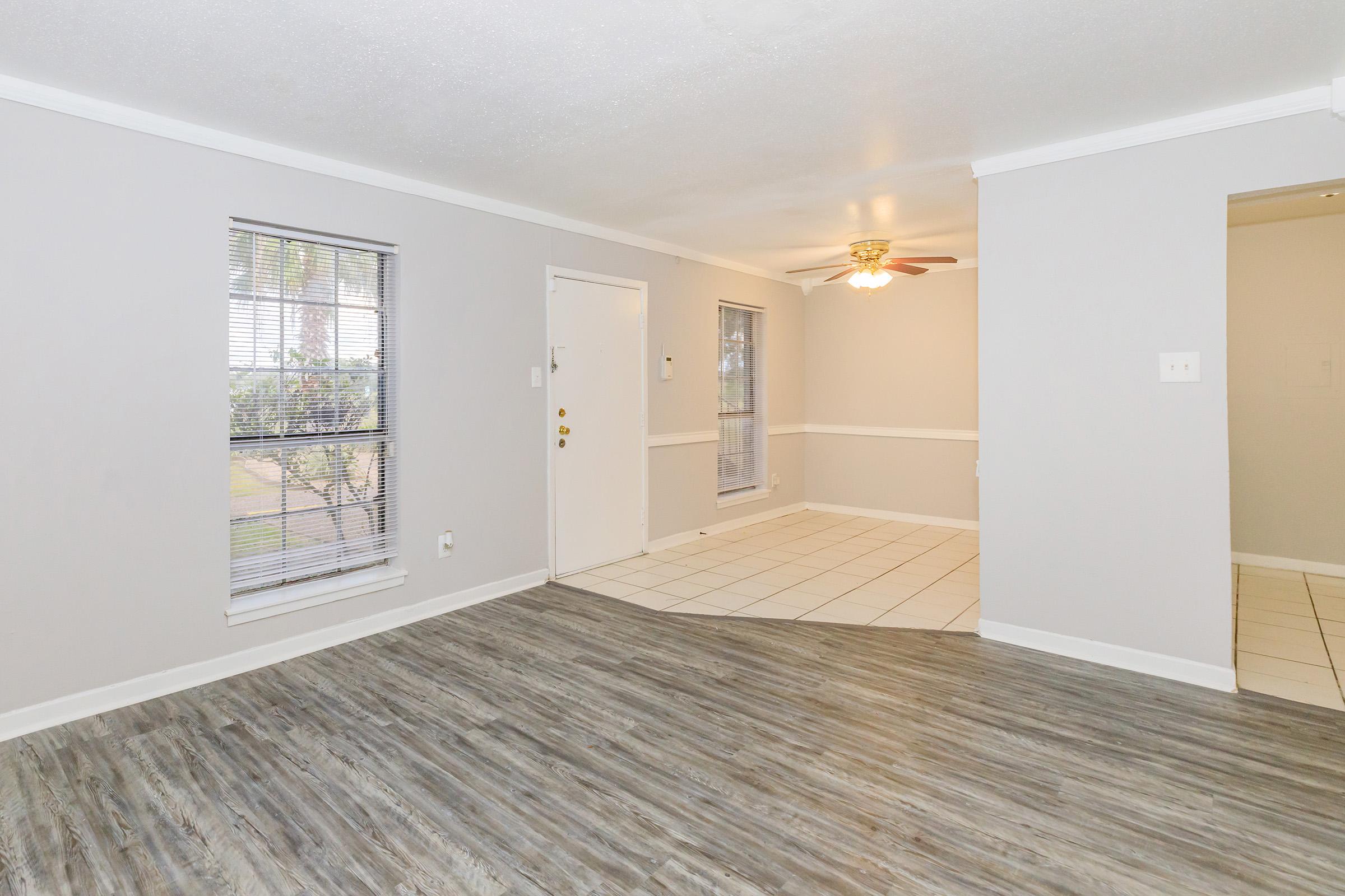 Bright and spacious living area featuring light gray walls, large windows with blinds, and a ceiling fan. The flooring is a mix of tiles and wood-like laminate. There’s a doorway leading to what appears to be a kitchen or dining area, creating an open layout.