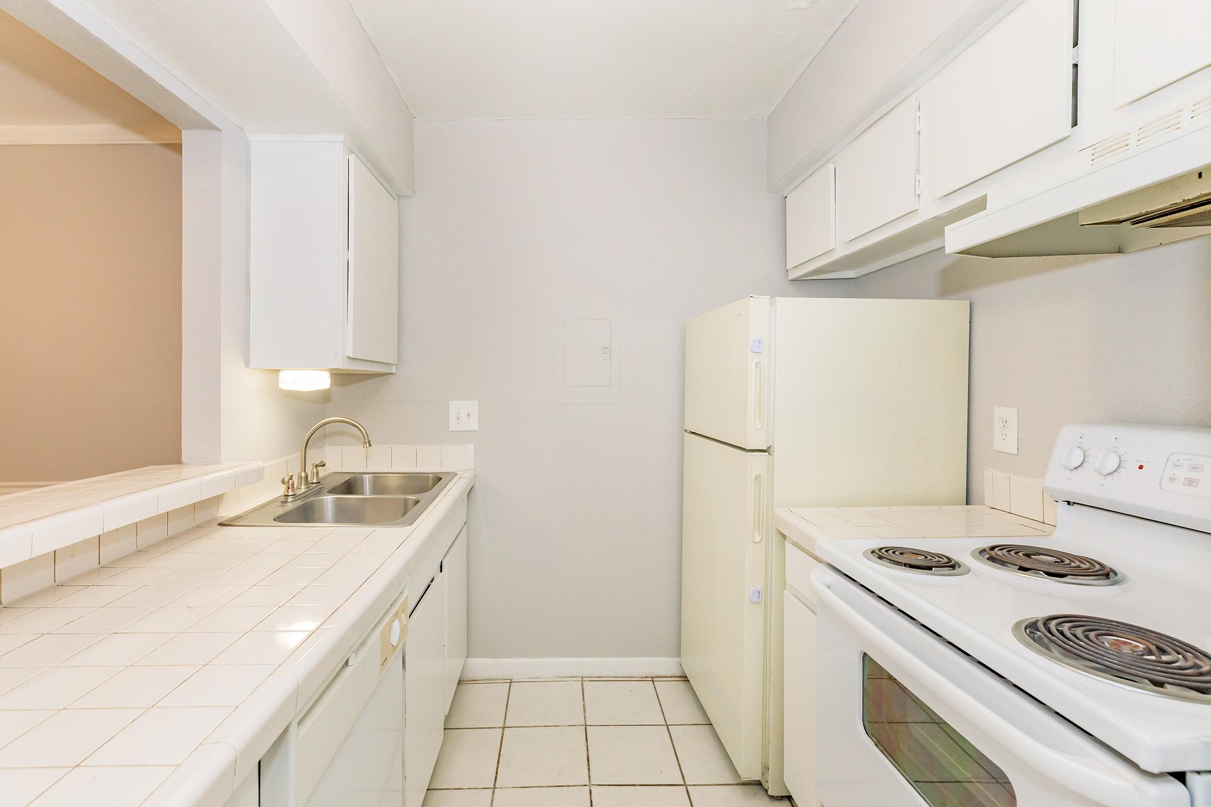 A clean, modern kitchen featuring white cabinetry, a double sink, a white refrigerator, and a white stove with an oven. The countertops are tiled, and the floor has square tiles. The walls are painted a light gray, creating a bright and airy atmosphere.