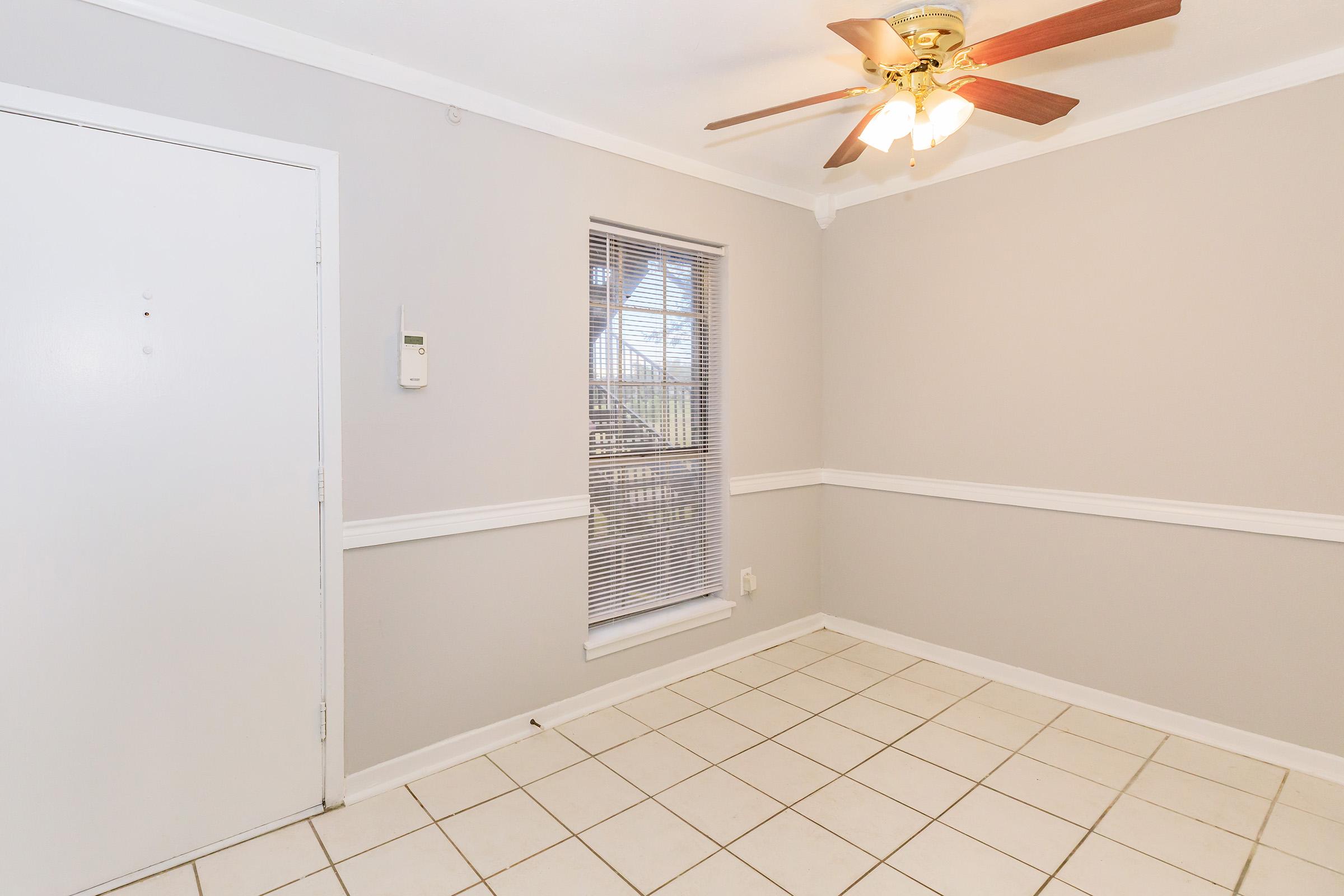 A light beige room with a ceiling fan, white trim, and beige tile flooring. There is a window with blinds and a white door on the left. The space is empty, creating a bright and open atmosphere.