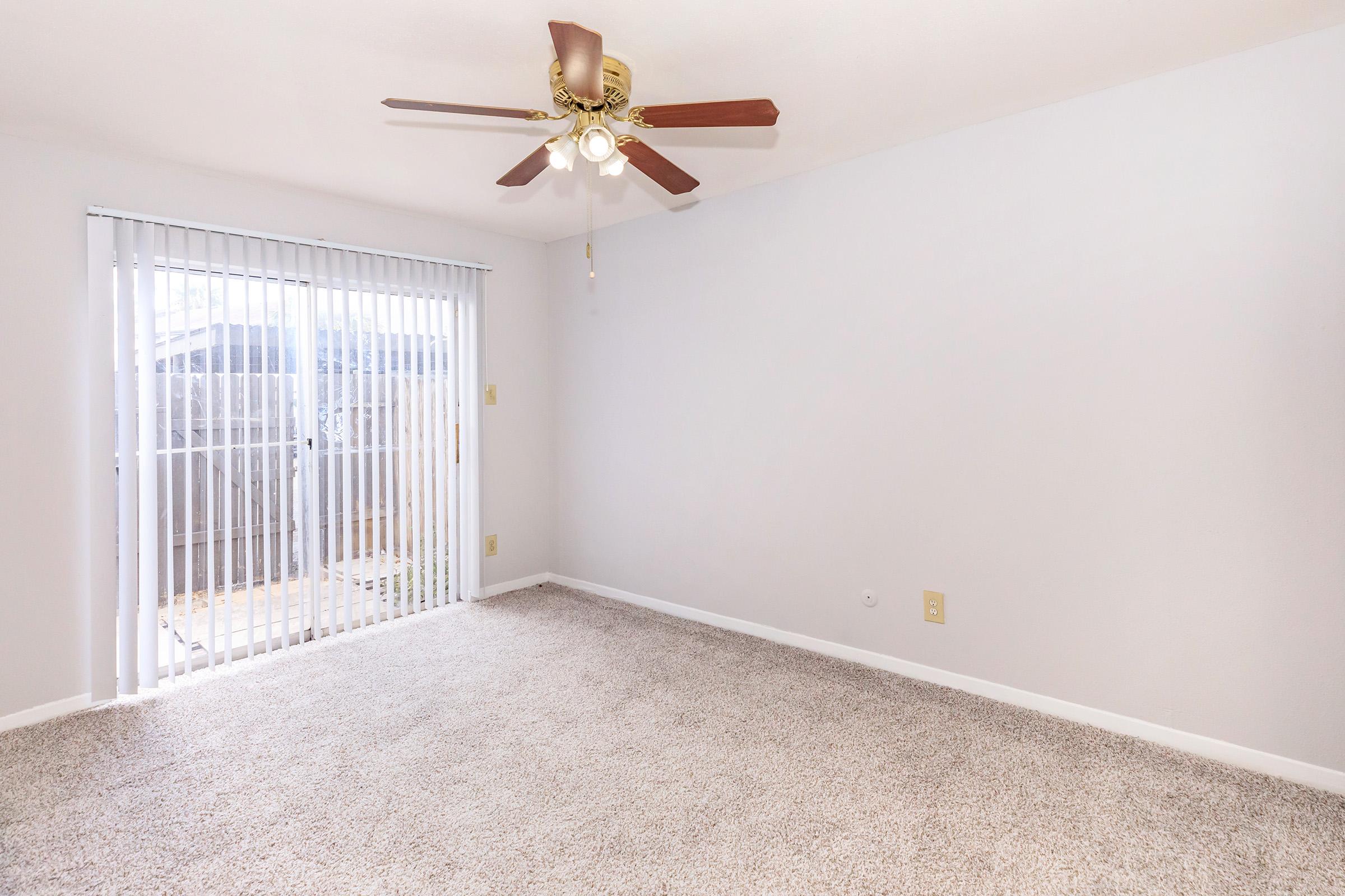 Empty room with light gray walls and a ceiling fan. The floor is covered with beige carpet, and there are vertical blinds covering a sliding glass door that leads to an outdoor patio area. Natural light is filtering in through the blinds, creating a bright, airy atmosphere.