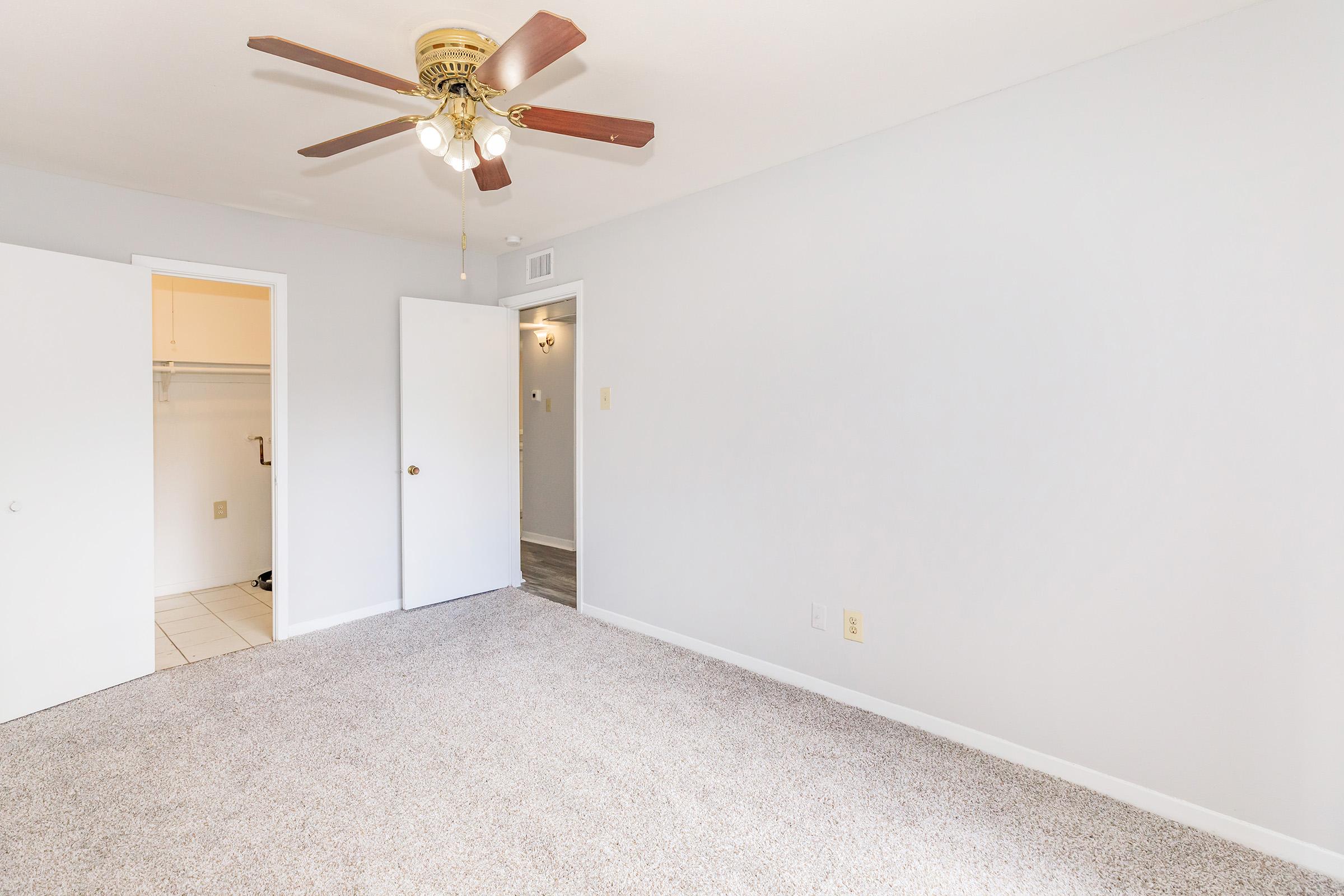 A spacious, well-lit bedroom featuring neutral-colored walls and plush carpet. There is a ceiling fan with wooden blades and light fixtures. One side has an open closet with sliding doors, while the opposite wall showcases a second door, leading to a bathroom or hallway.
