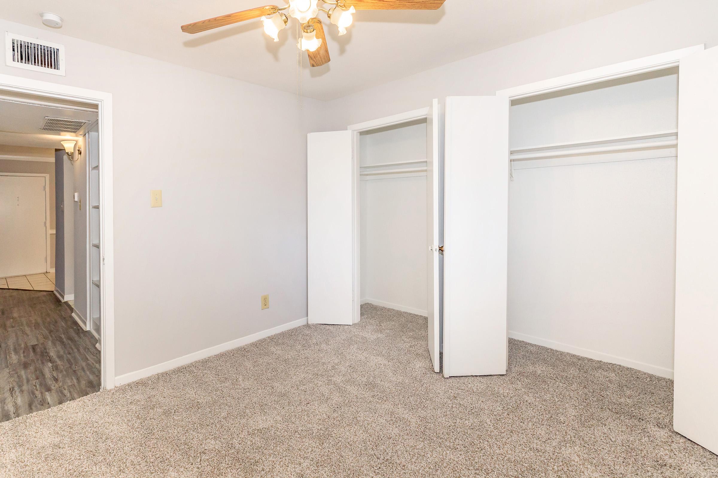 A well-lit bedroom with light-colored walls and a ceiling fan. The room features two white sliding closet doors on the right and a plush, carpeted floor. The doorway to another room is visible on the left, suggesting a spacious layout.