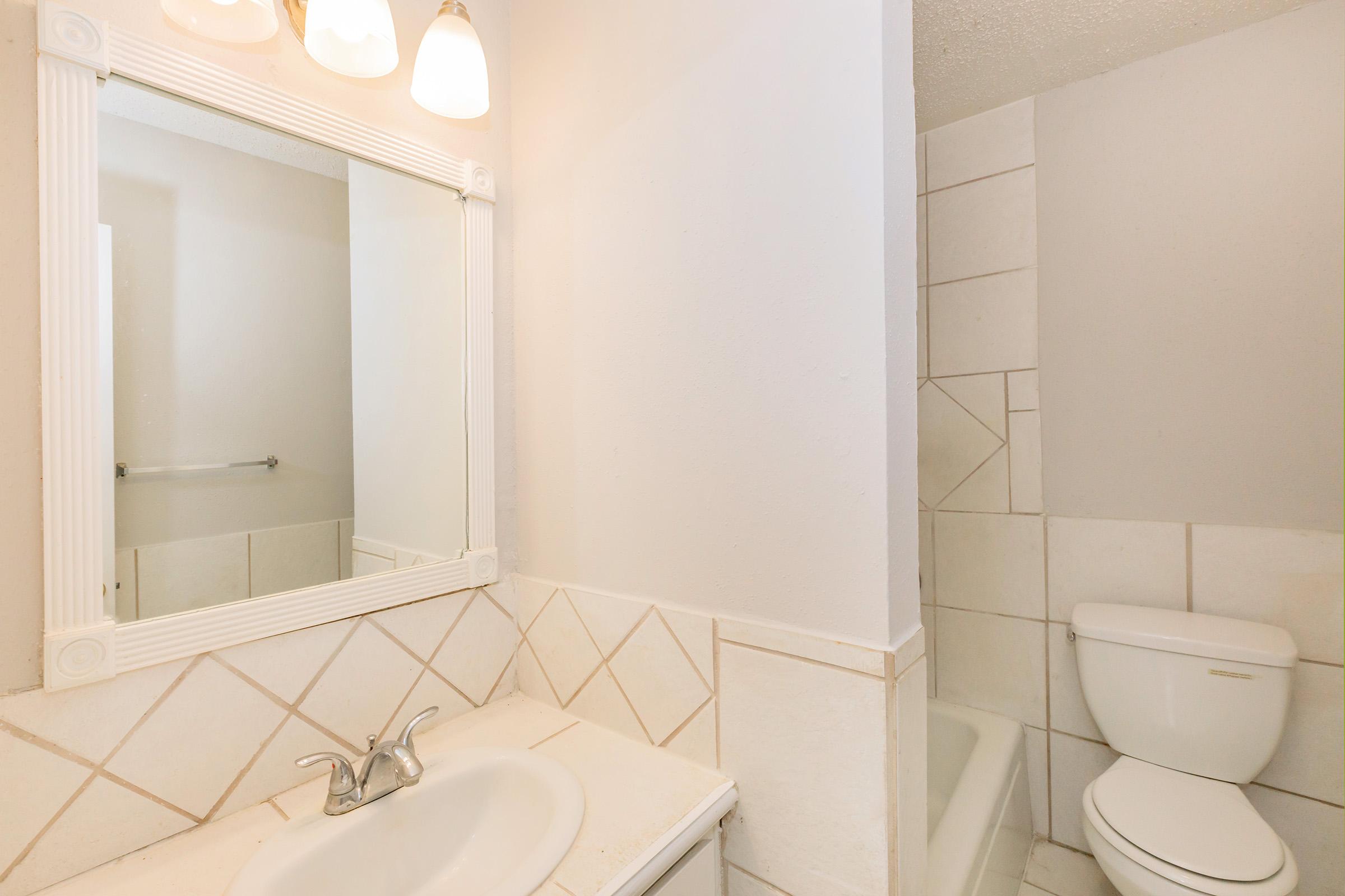A bright bathroom featuring a mirrored vanity with a sink and faucet, tiled backsplash, and a small shower area with white tiles. A toilet is positioned next to the shower. The walls are painted in a light color, enhancing the airy feel of the space.