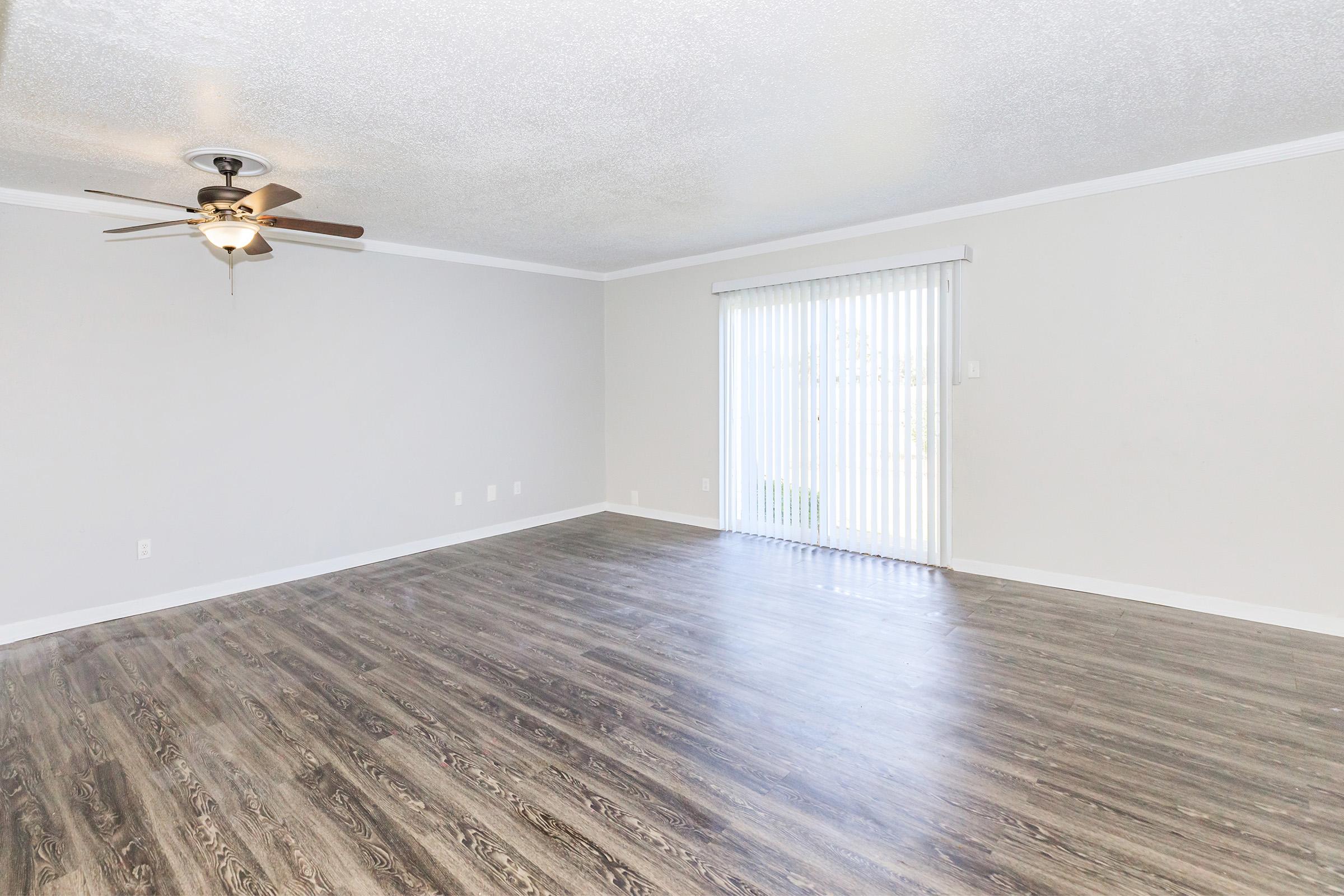 A spacious, empty living room featuring light-colored walls, wooden flooring, and a ceiling fan. A large window with vertical blinds allows natural light to enter, creating a bright and airy atmosphere. The room is unadorned and ready for furniture arrangement.