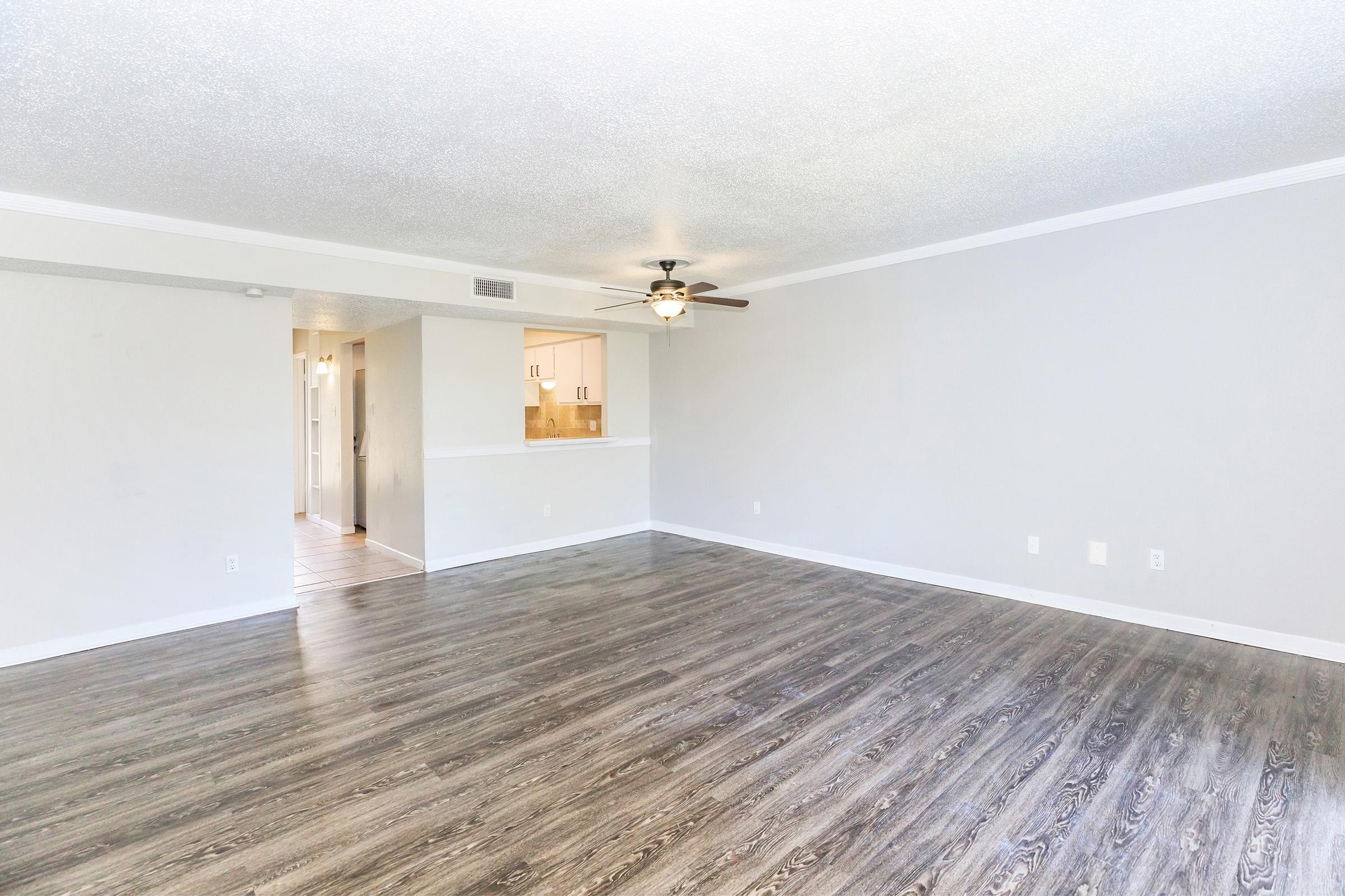A spacious, empty living room featuring a light-colored wall, hardwood-style flooring, and a ceiling fan. A doorway in the background leads to another room, and there is a subtle division with a short wall where a kitchen or dining area is visible. Natural light brightens the space.