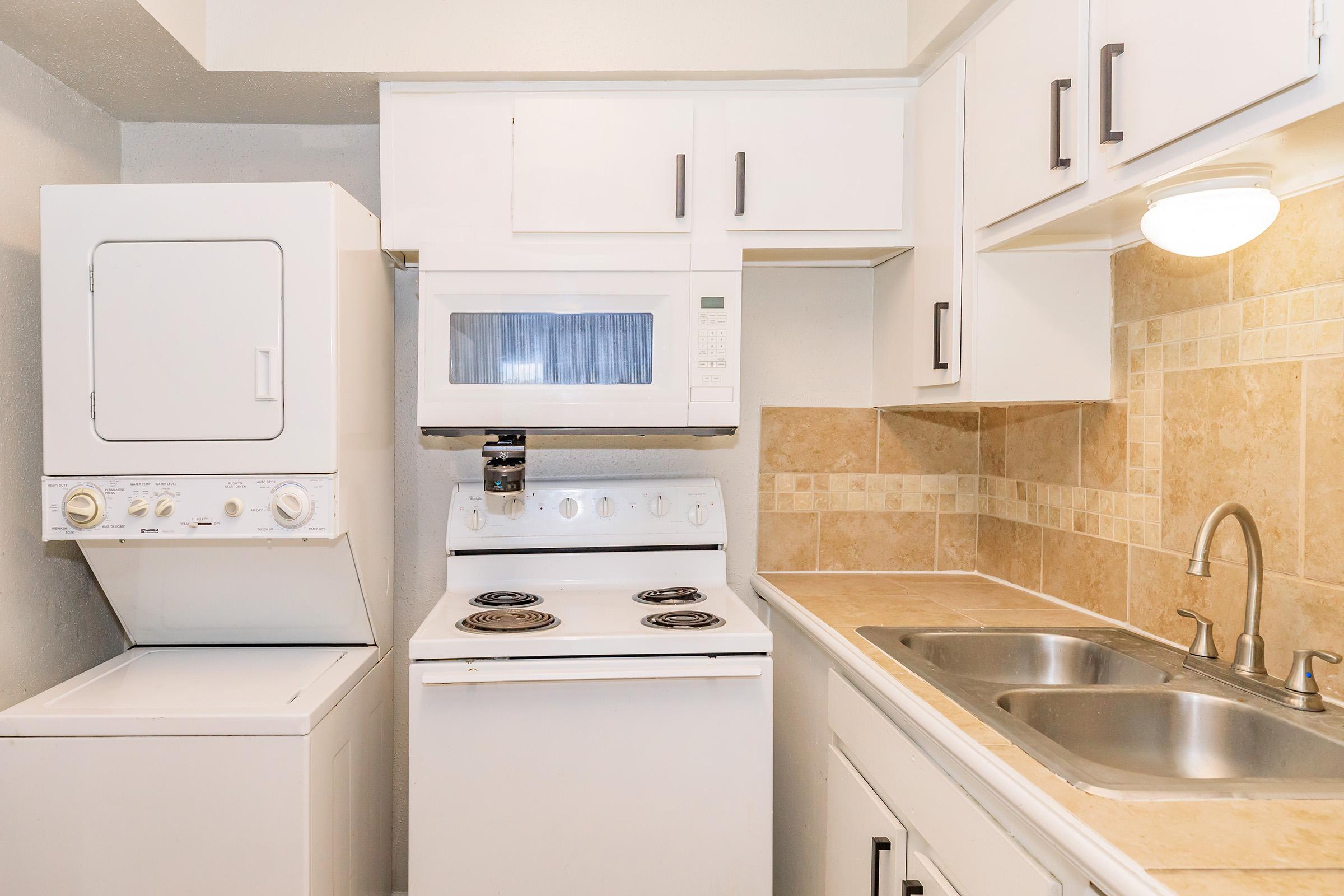 A small, modern kitchen featuring a stacked washer and dryer on the left, a microwave and stove in the center, and a double sink on the right. The kitchen has light-colored cabinets and a tiled backsplash, creating a clean and functional space.