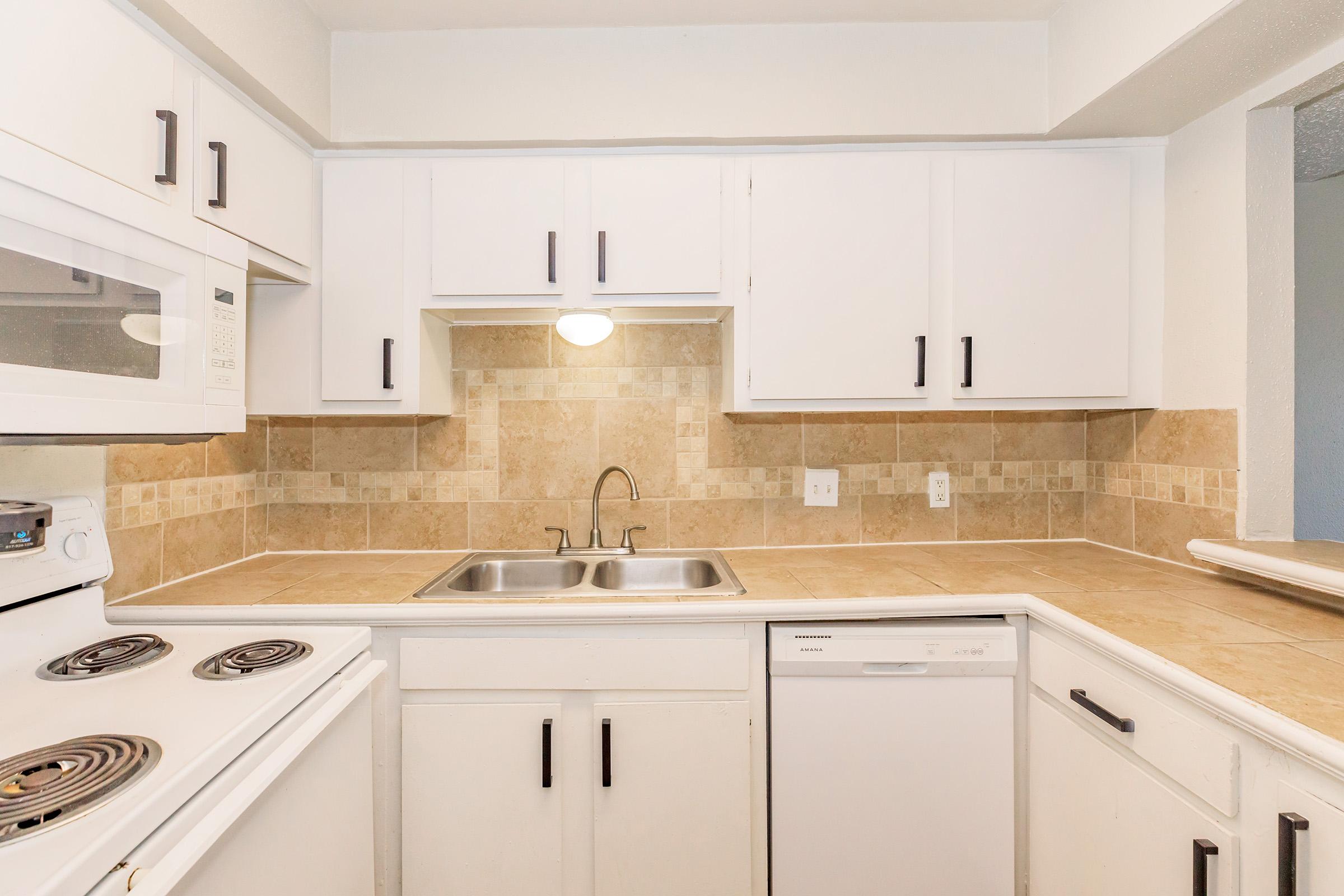 A modern kitchen with white cabinetry, beige tile backsplash, stainless steel sink, and countertop. Appliances include a white dishwasher, a stovetop with burners, and a microwave. The space is well-lit, creating a clean and functional cooking area.