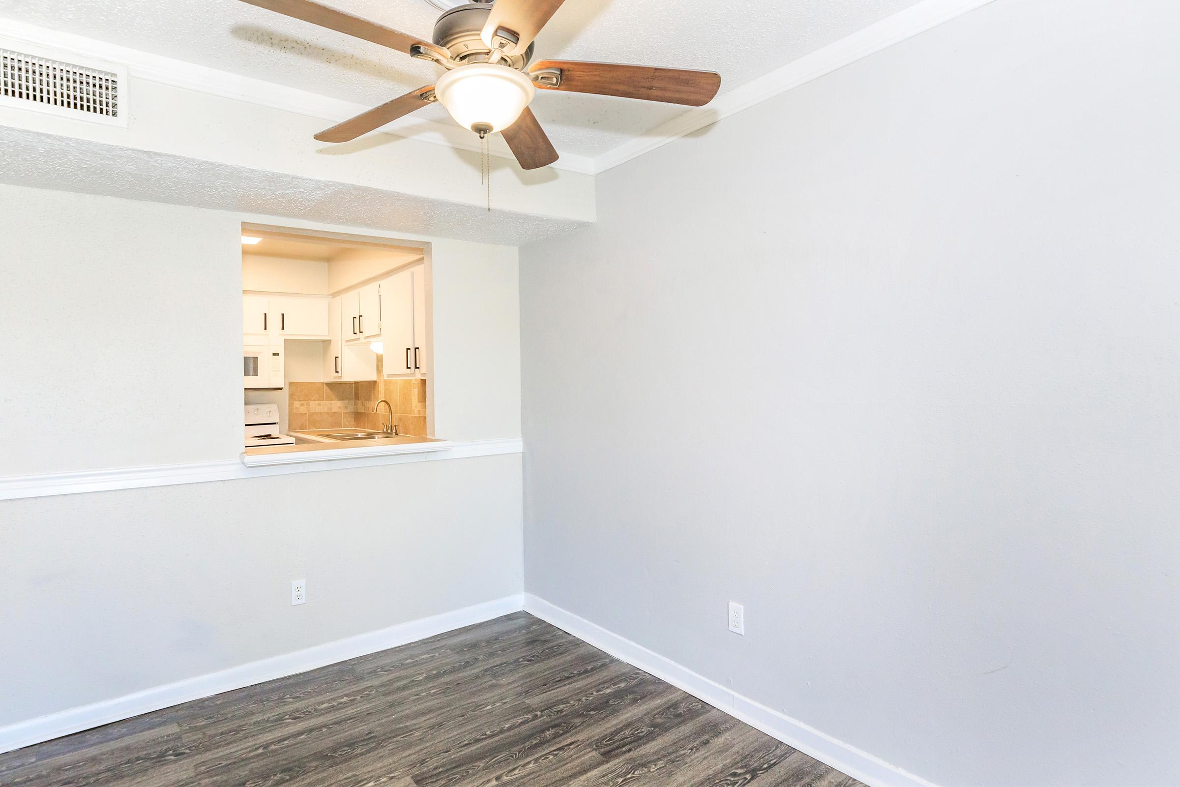 A small, empty room with light gray walls and a ceiling fan. There's a window with an open view into a kitchen area featuring white cabinets. The floor is covered in wood-like laminate. The room appears bright and well-lit, giving a spacious feel despite its compact size.