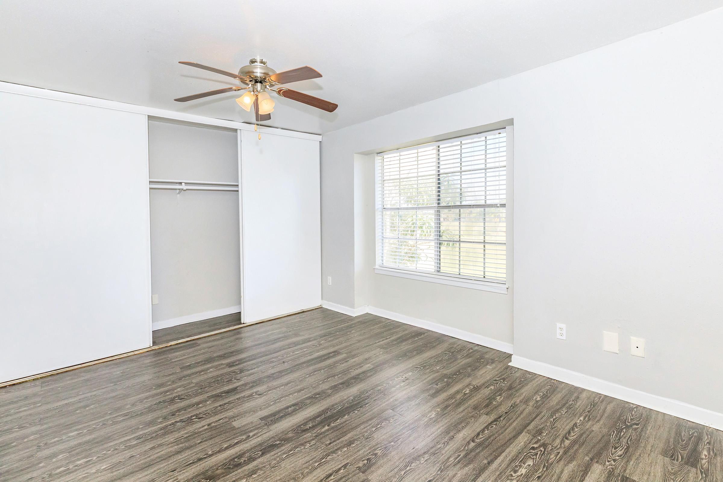 A bright, empty bedroom featuring light gray walls and a ceiling fan. It has a large window with blinds, allowing natural light to enter, and wooden-looking laminate flooring. There is a closet with sliding doors on one side, providing storage space.