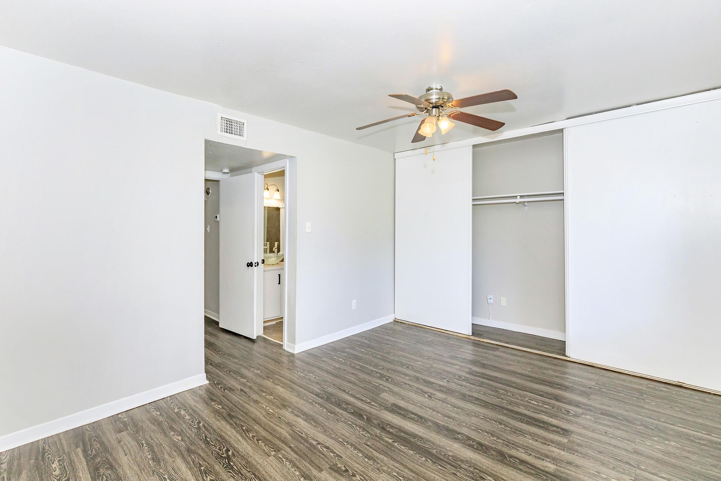A spacious room featuring light-colored walls, a ceiling fan with wooden blades, and laminate flooring. There's a closet to the right and a doorway leading to another area. Natural light brightens the room, creating a welcoming atmosphere.