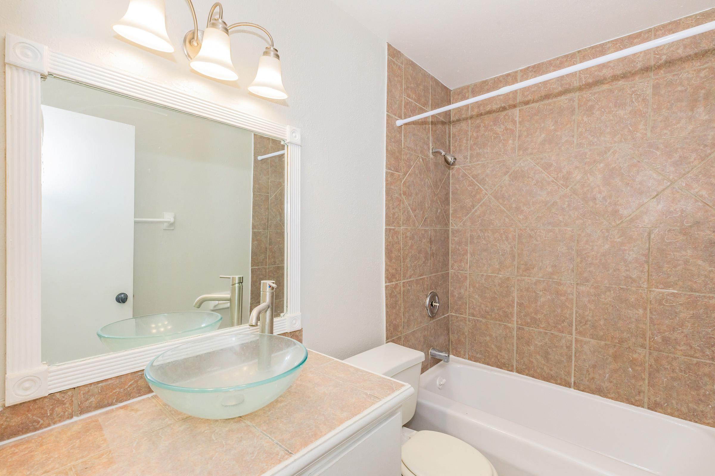 Modern bathroom featuring a glass vessel sink atop a beige countertop, a wall-mounted mirror with three light fixtures, and a bathtub with a shower. The walls are covered in earthy-toned tiles, creating a warm and inviting atmosphere. A towel bar is visible, and the room is well-lit.
