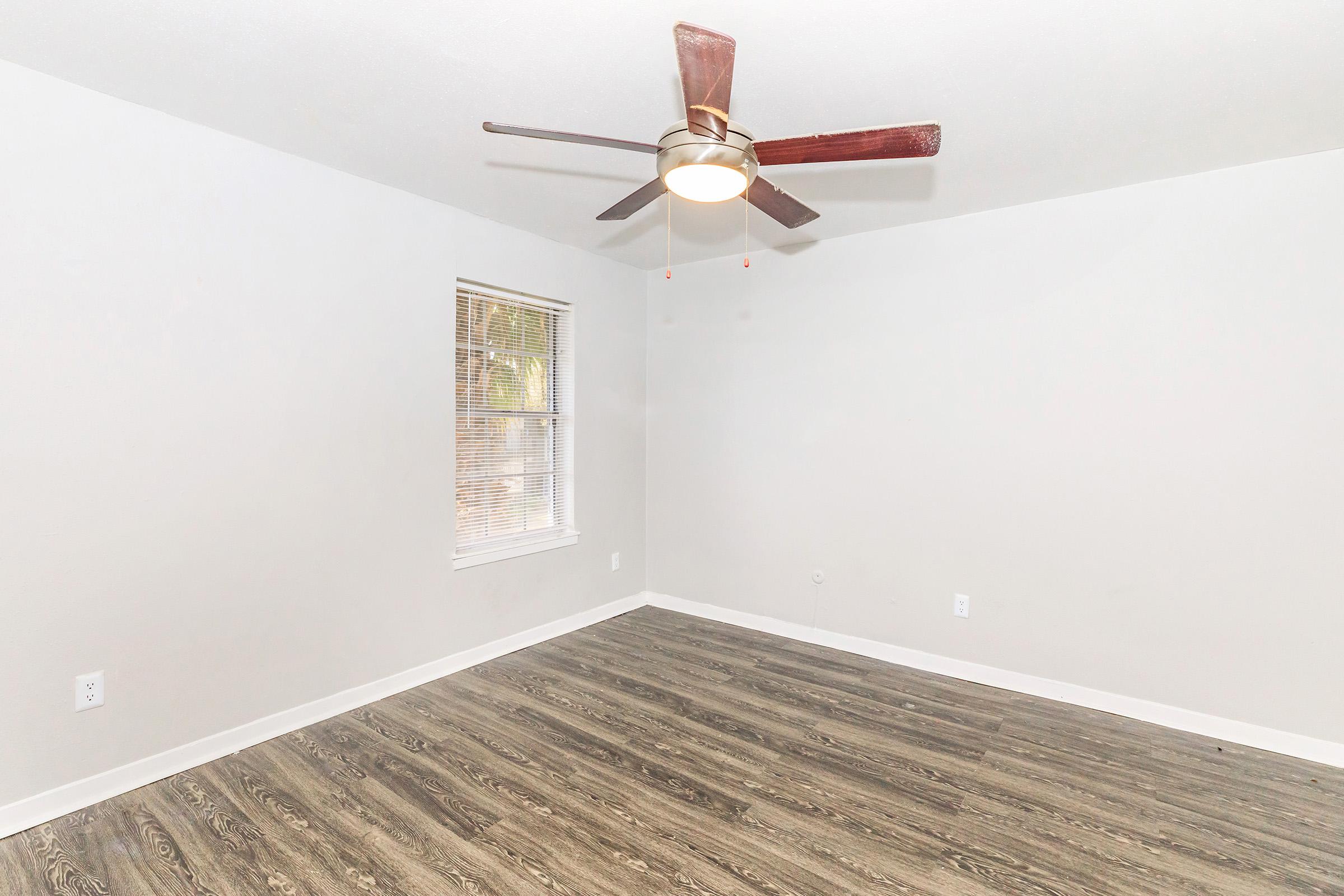 A spacious room with light gray walls and a ceiling fan. It features a laminate floor that resembles wood and a large window with blinds, allowing natural light to enter. The space is empty and ready for furnishing.