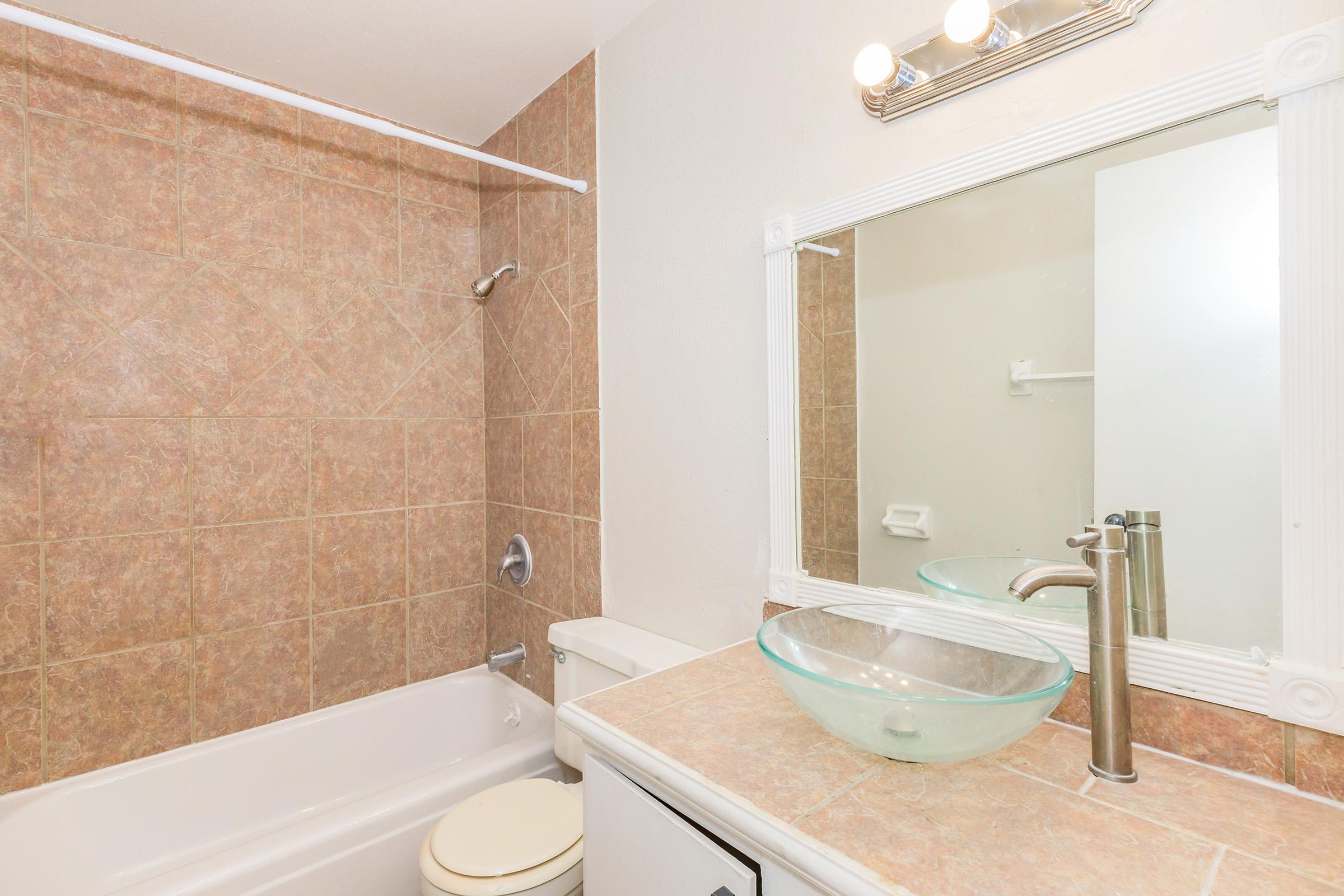 A modern bathroom featuring a bathtub with a showerhead, a glass vessel sink on a countertop, and a large mirror above the sink. The walls and tub surround are tiled in a light brown pattern. The bathroom has bright lighting and a clean, minimalist design.
