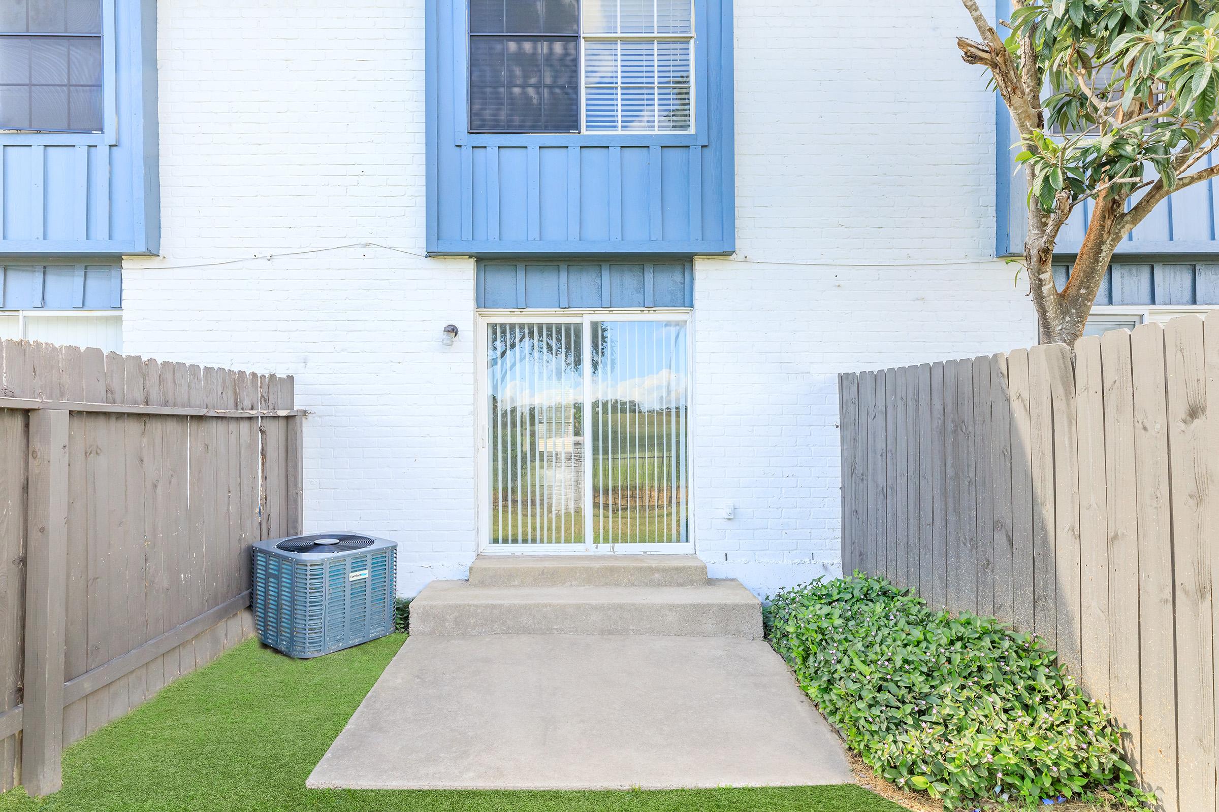 A small outdoor patio area with a concrete step leading up to a glass sliding door. The patio is enclosed by a wooden fence, with an air conditioning unit on the left and lush green plants bordering the space, set against a light-colored brick wall.