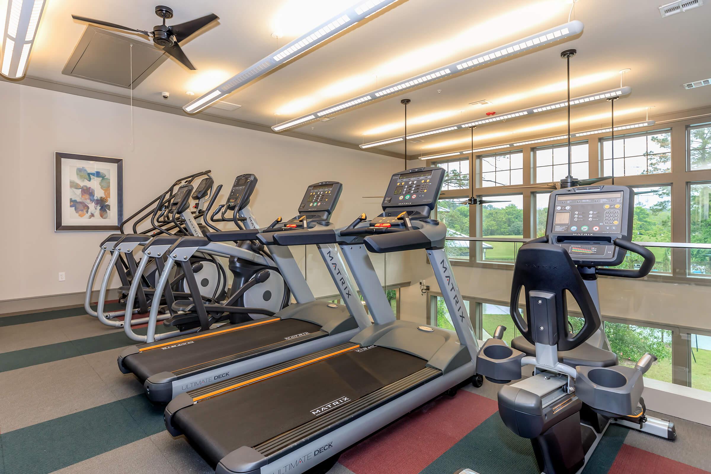 Treadmills and cycles on second story of fitness center facing large windows.