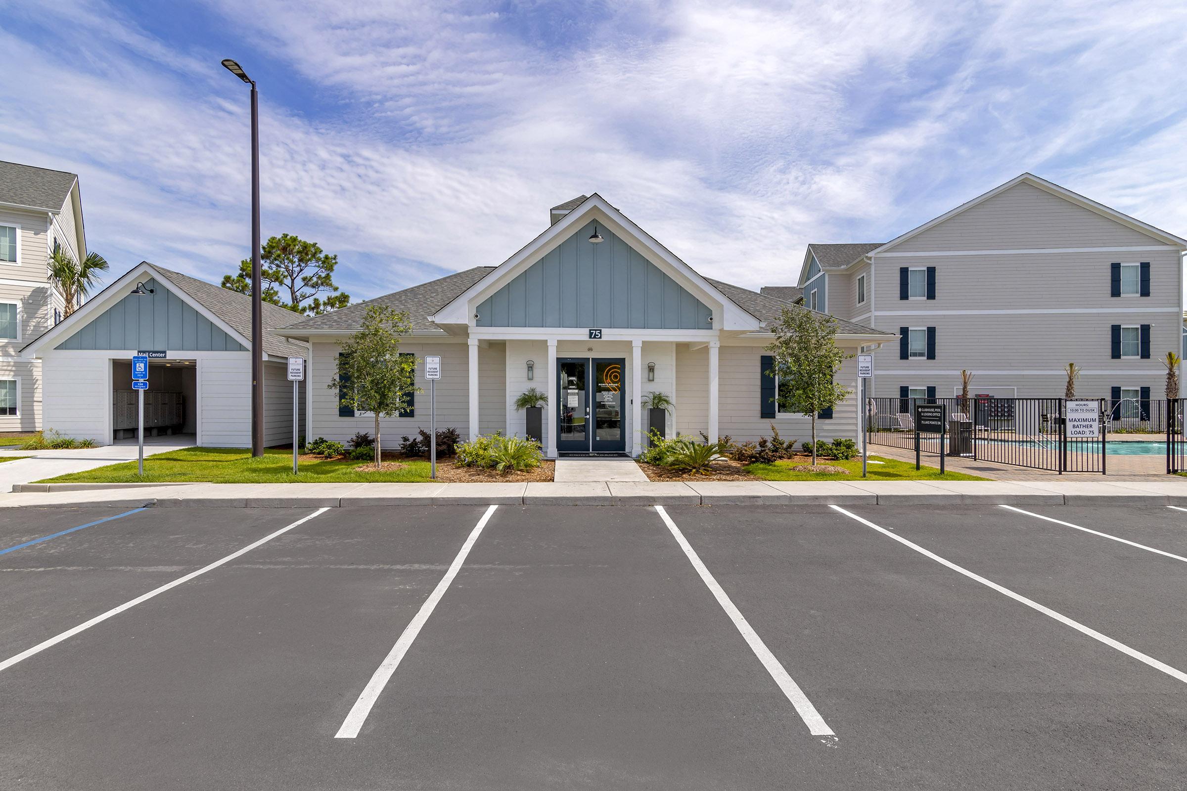 A modern building with a light blue exterior, featuring a covered entrance flanked by small trees. Nearby, there are designated parking spaces and a pool area visible behind a fence. The scene is set under a clear blue sky with some clouds, suggesting a pleasant, sunny day.