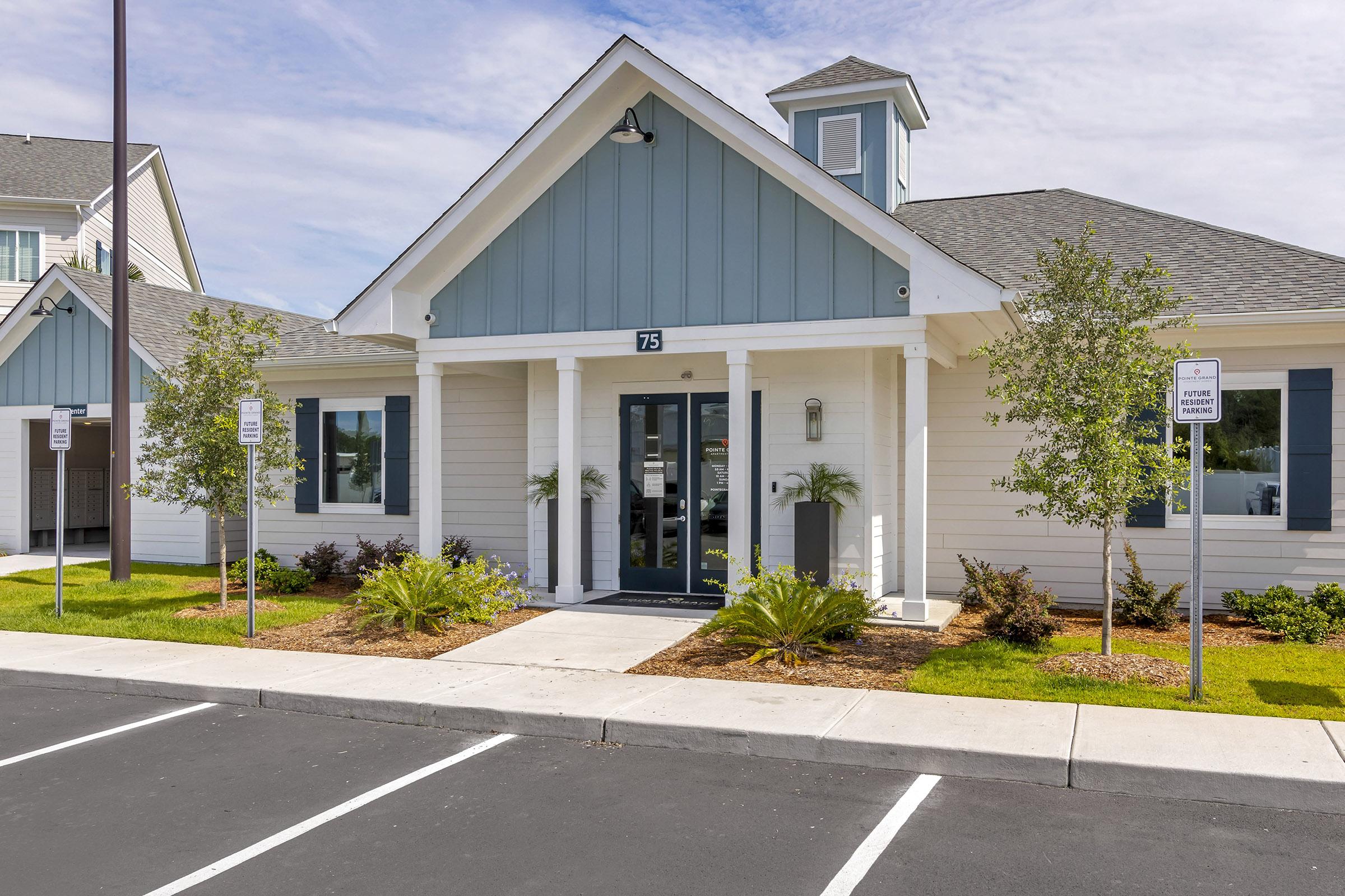 A modern building with a light blue and white exterior, featuring a sloped roof and a small tower. The entrance has a large glass door, flanked by greenery and shrubs. There are parking spaces visible in front, and the area is well-maintained, surrounded by grass and landscaping.
