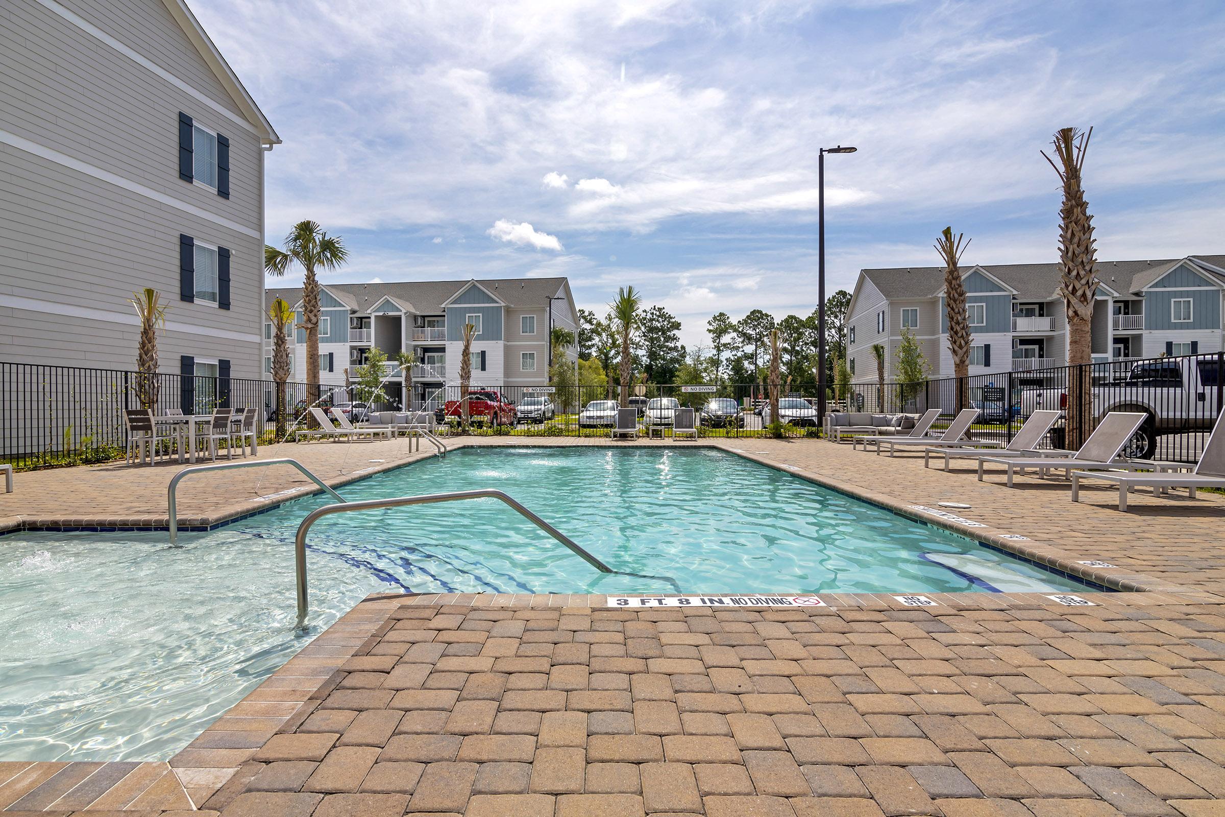 A clear swimming pool surrounded by lounge chairs and palm trees, located within a residential area. Apartments are visible in the background, with a blue sky and fluffy clouds above. The pool area features tiled decking and safety signage.