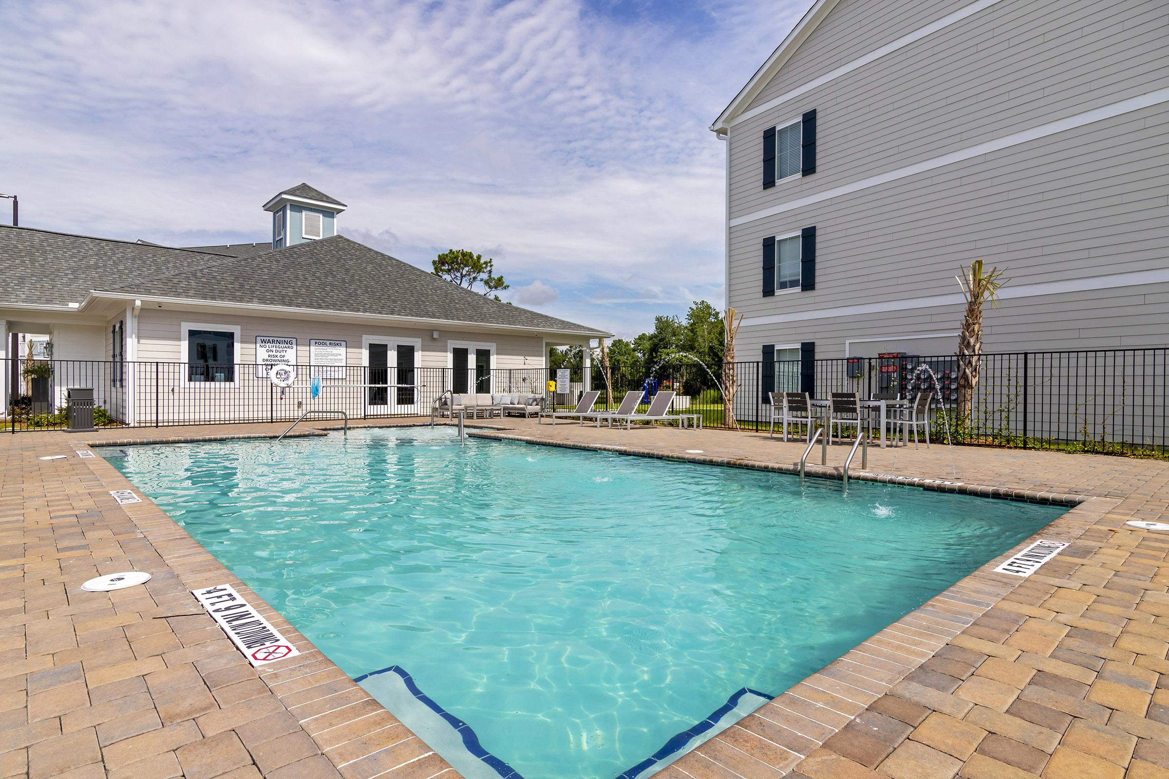 A clean, inviting outdoor swimming pool surrounded by a brick patio. There are lounge chairs nearby, and a building with large windows and a peaked roof is in the background. The sky is partly cloudy, adding to the serene ambiance of the area.