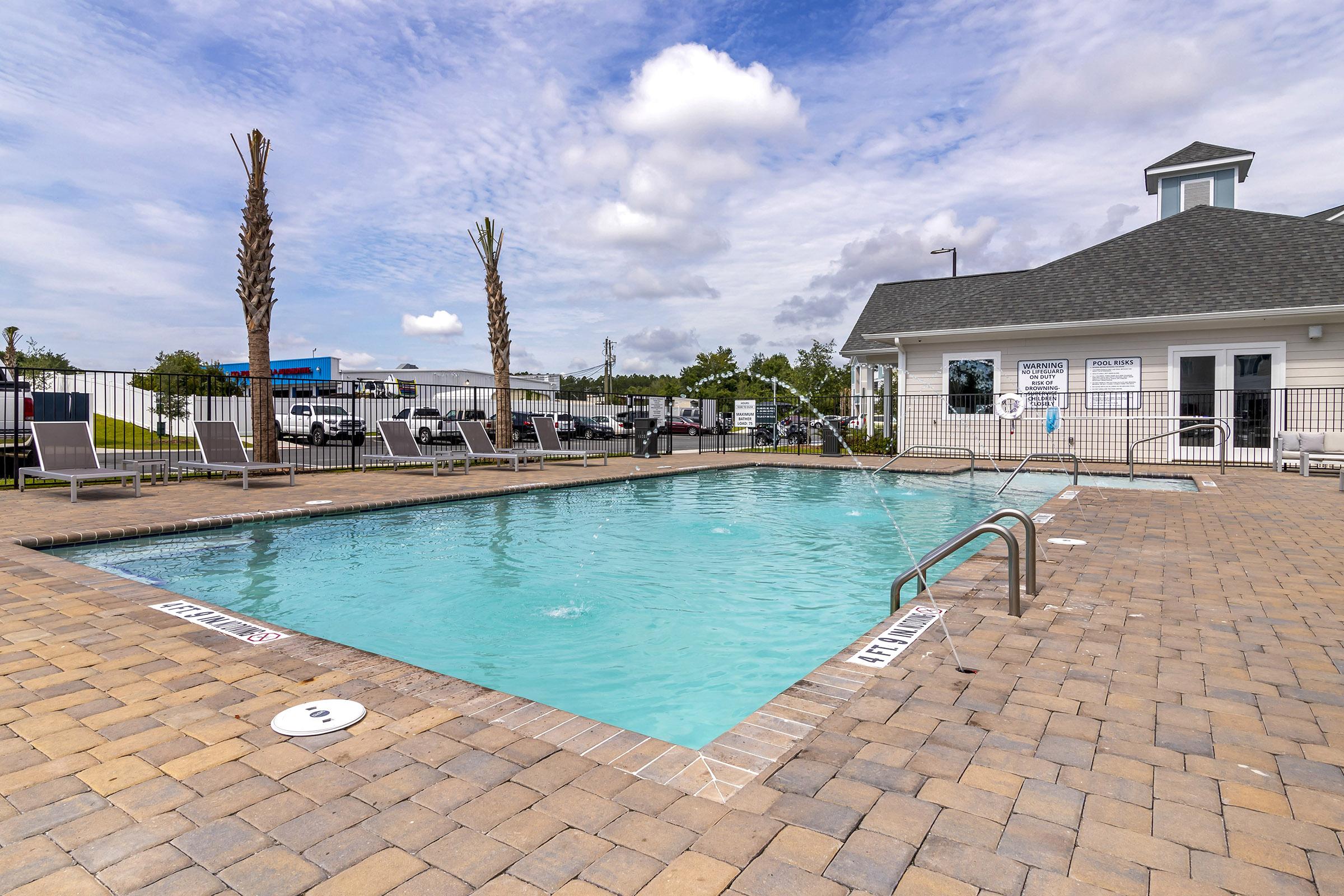 A clear outdoor swimming pool surrounded by a patio of light-colored pavers. Palm trees line one side, and there are lounge chairs in the background. A building with a gray roof is visible next to the pool, and the sky is partly cloudy.