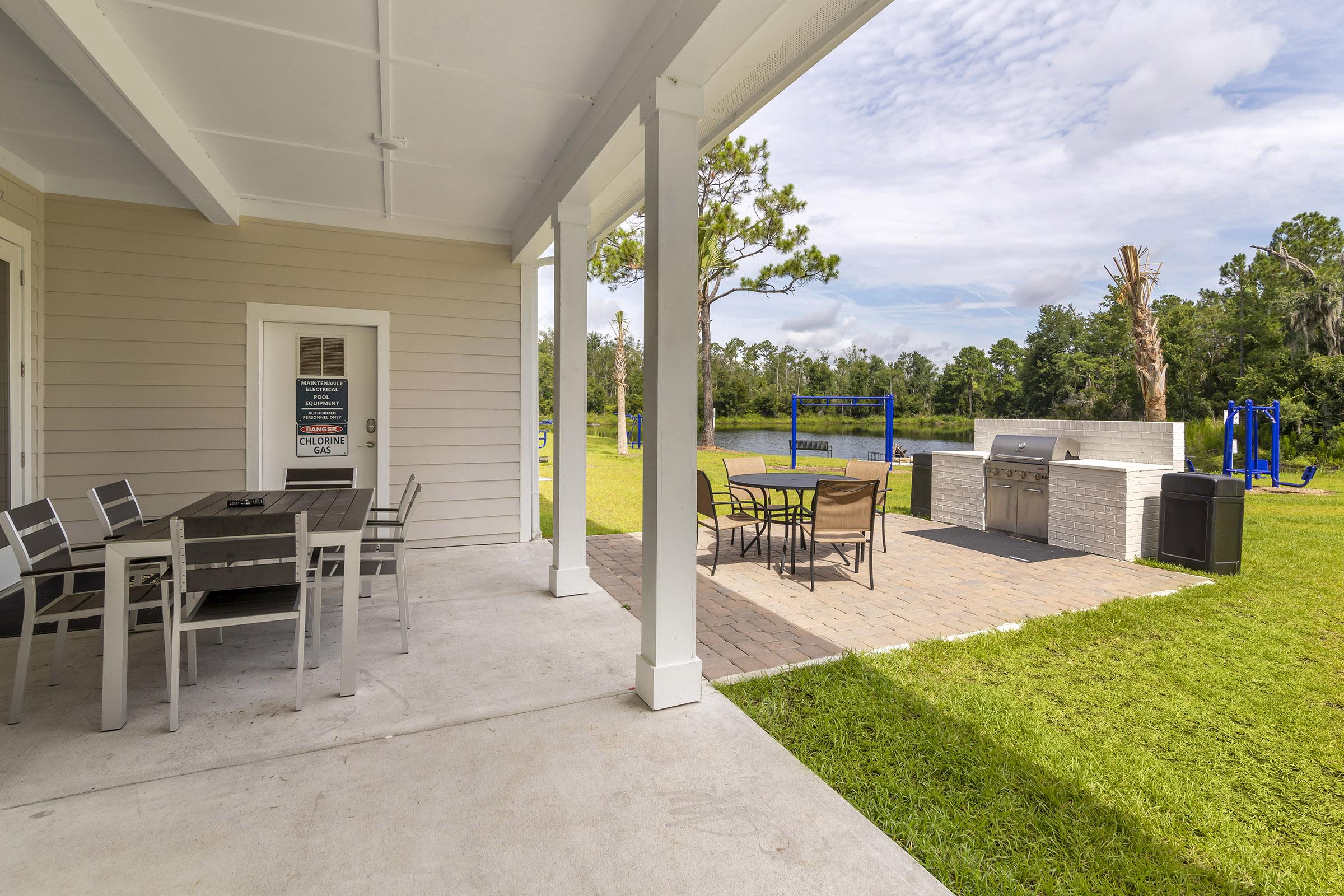 A backyard patio with a dining table and chairs, an outdoor grill area, and grass surrounding the space. In the background, there are blue swings and a swing set, with trees and a cloudy sky visible. The setting appears spacious and ideal for outdoor gatherings.