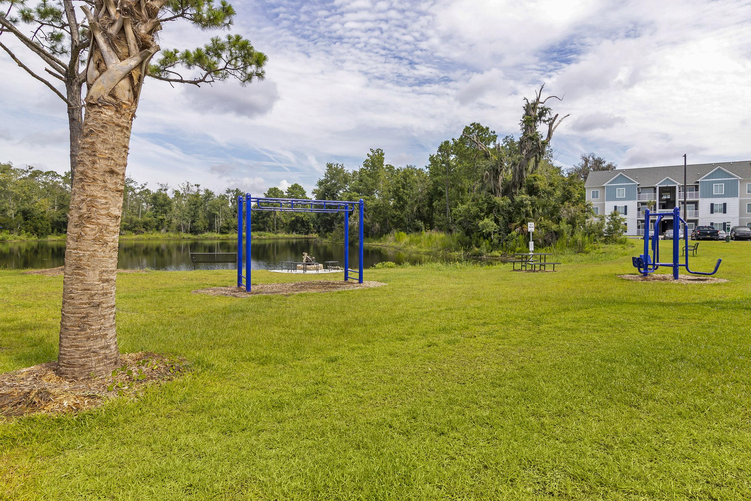 A grassy area by a pond features two blue swings and fitness equipment. In the background, there are trees and a residential building. The sky is partly cloudy, creating a serene outdoor setting ideal for recreation and relaxation.