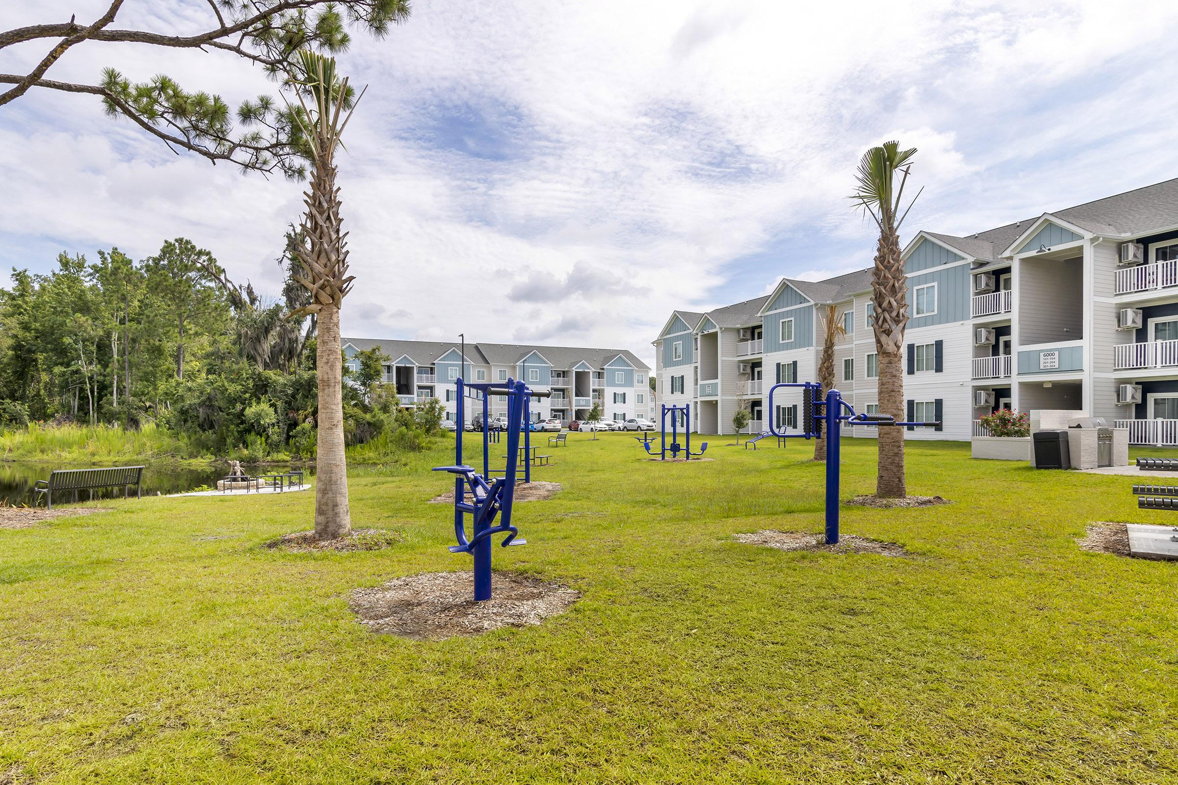 Outdoor fitness area with exercise equipment on grassy lawn, surrounded by palm trees. In the background, there are multi-story residential buildings with balconies, set against a cloudy sky and green foliage. The scene is tranquil and inviting for outdoor workouts.