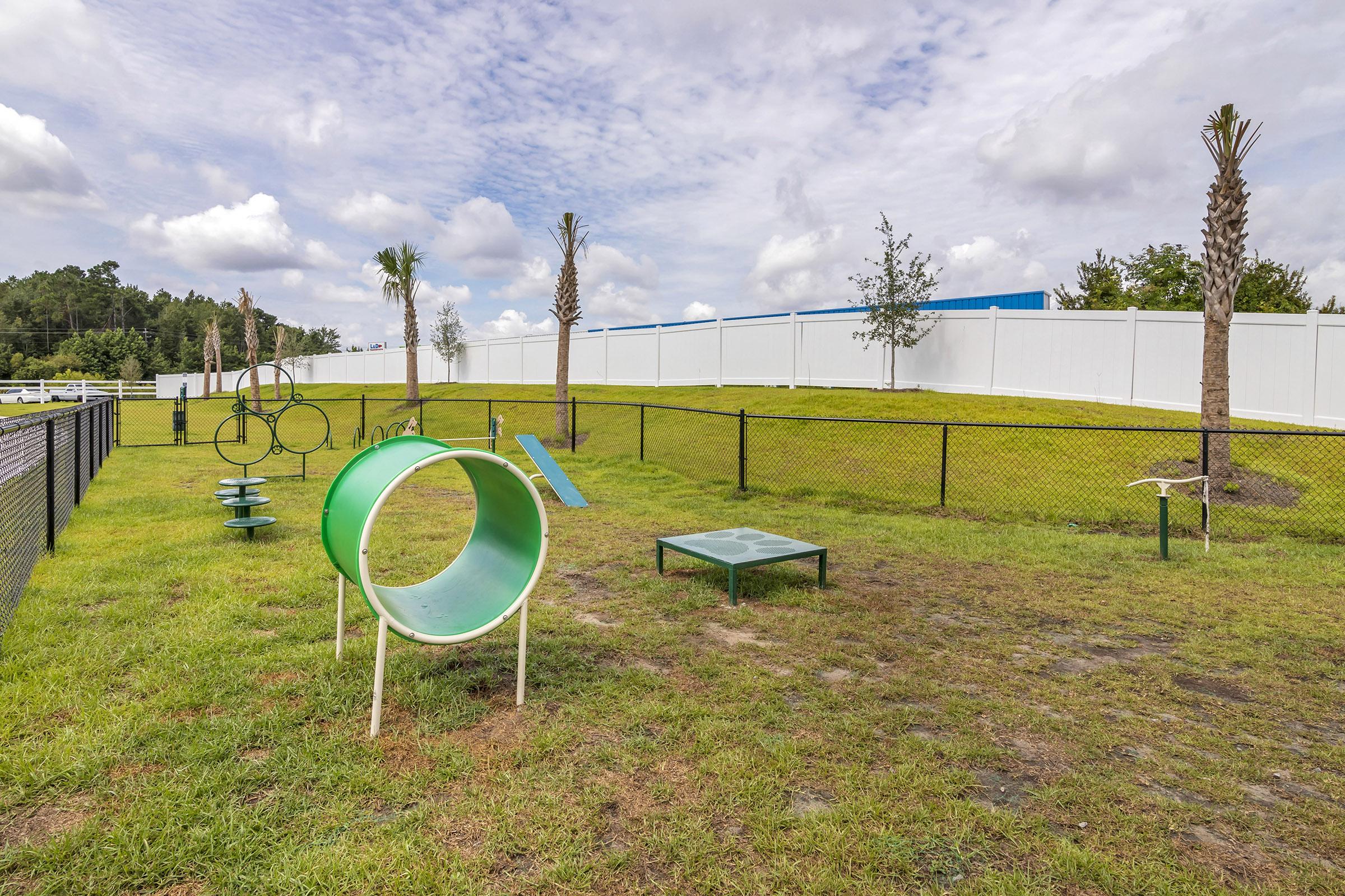 A grassy area in a fenced play space featuring various playground equipment, including a tunnel, climbing structures, and balance beams. The background includes a white fence and palm trees under a partly cloudy sky.