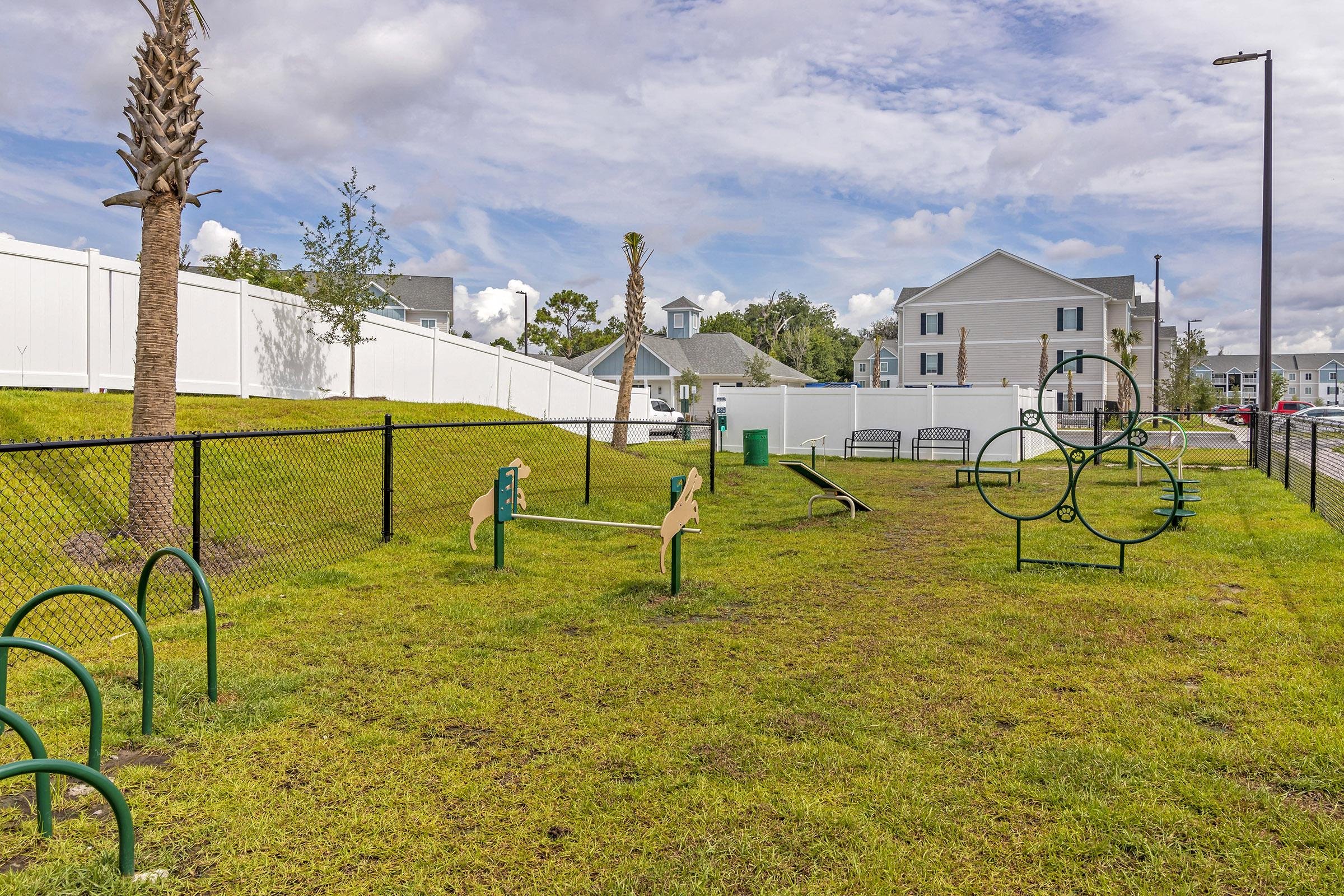A grassy dog park enclosed by a black fence, featuring agility equipment like a seesaw, hoops, and climbing structures. Palm trees are visible in the background, along with residential buildings and a cloudy sky.