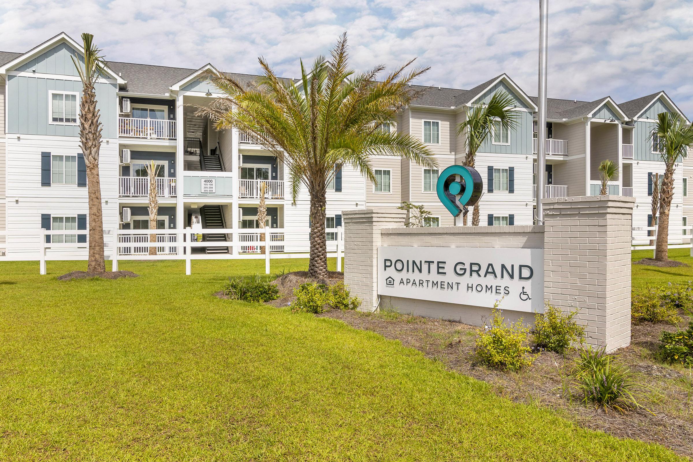 Exterior view of Pointe Grand Apartment Homes, showcasing a well-maintained building with light-colored siding, balconies, and palm trees in the foreground. The entrance features a sign with the apartment name, set against a green lawn under a partly cloudy sky.