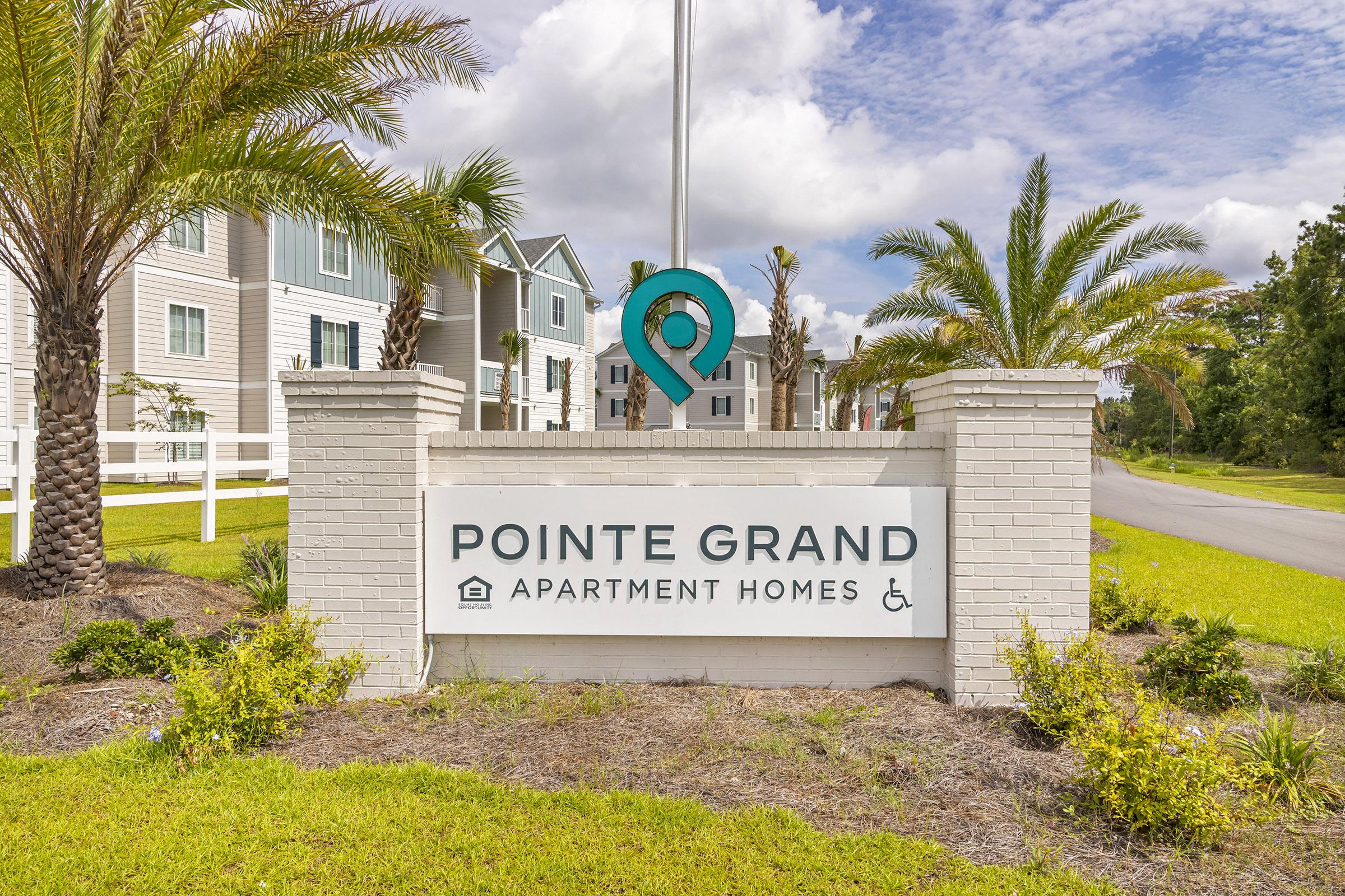 Sign for Pointe Grand Apartment Homes featuring a blue location pin emblem, surrounded by well-maintained landscaping with palm trees and grass. A clear blue sky overhead with a few clouds, emphasizing a welcoming atmosphere for potential residents.