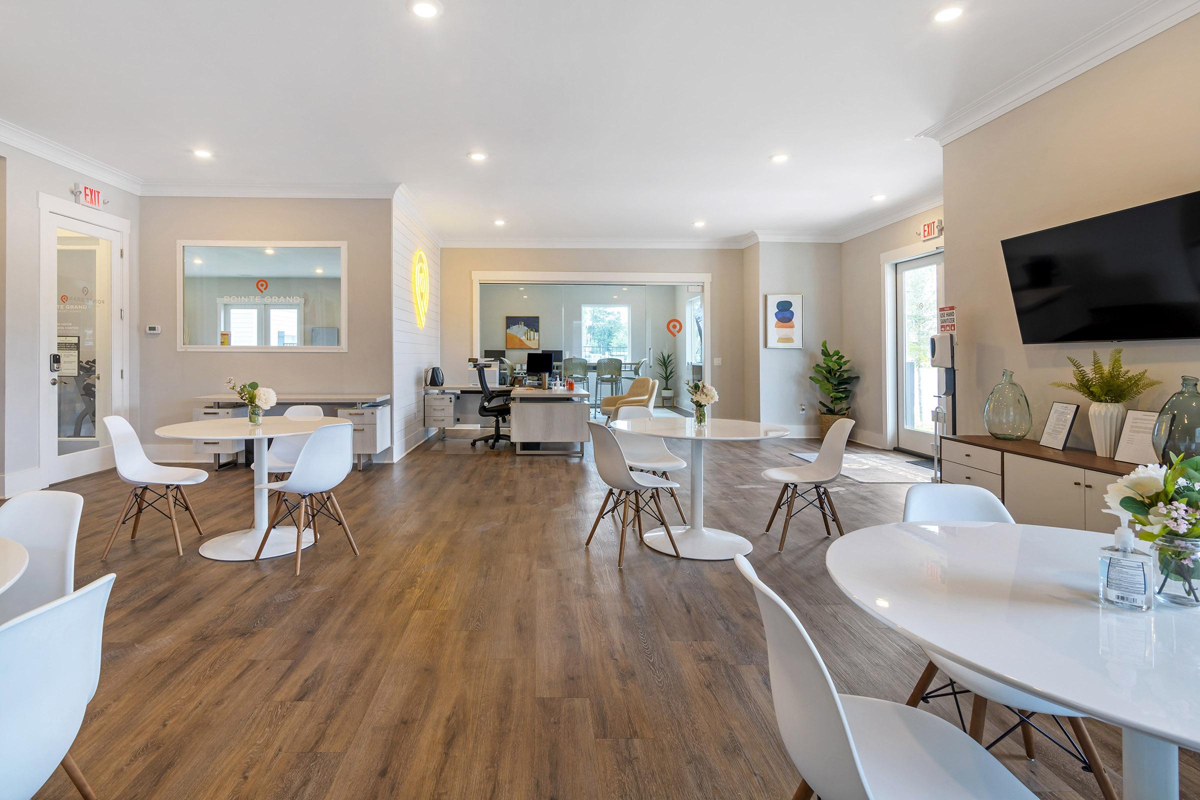 A modern office space featuring white round tables and minimalist chairs. The room has wooden flooring and is well-lit, with a reception area visible in the background. There are plants and decorative elements, creating a welcoming atmosphere for both clients and staff.