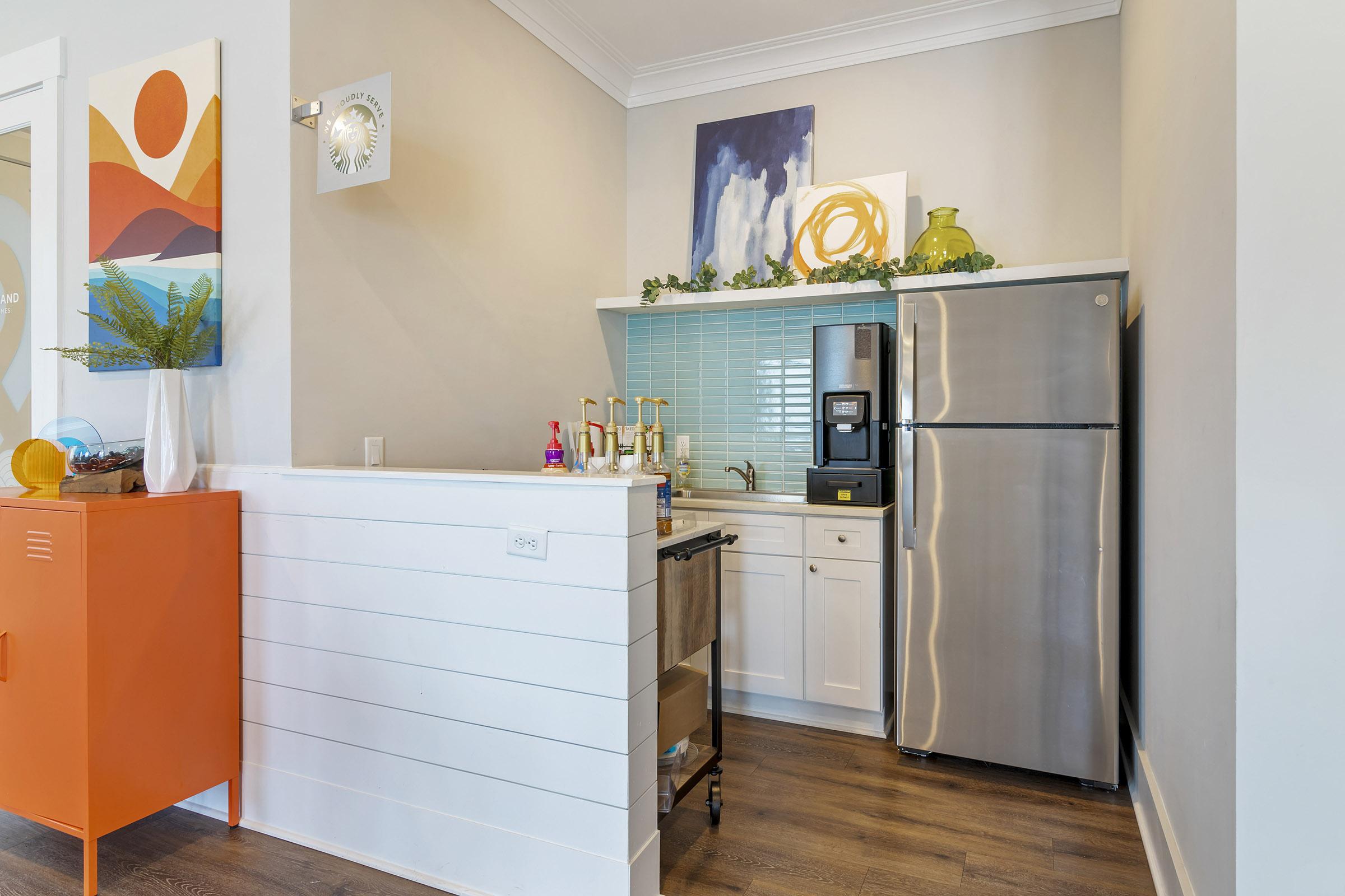 Contemporary kitchen featuring white cabinetry, a stainless steel refrigerator, and a stove. A light blue tile backsplash complements decorative elements, including green and orange accents. Artwork with abstract designs hangs on the wall, enhancing the modern aesthetic. Bright and inviting space with natural light.