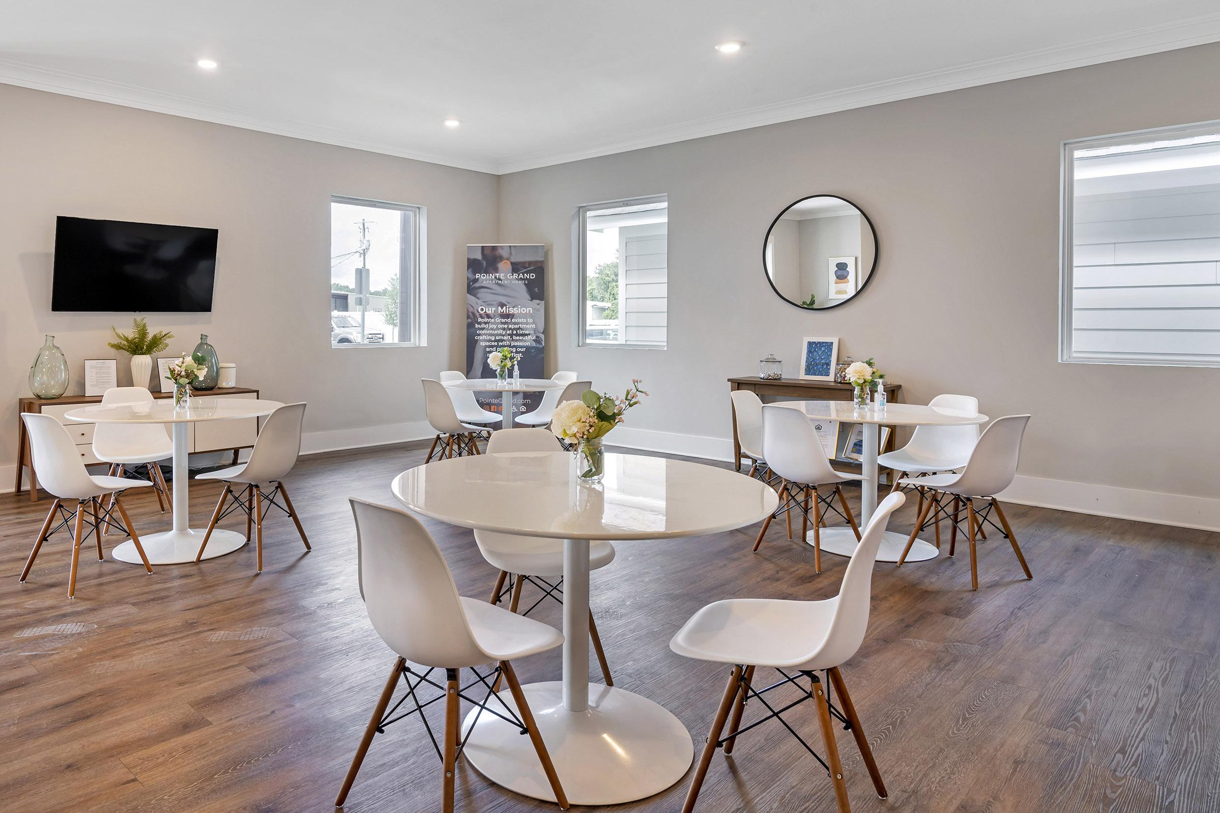 A modern, well-lit interior featuring several round tables with white chairs, arranged for dining or meetings. A large mirror is mounted on the light gray wall, and windows let in natural light. Decorative elements include small flower vases on the tables. A TV is mounted on the wall, adding to the ambiance.